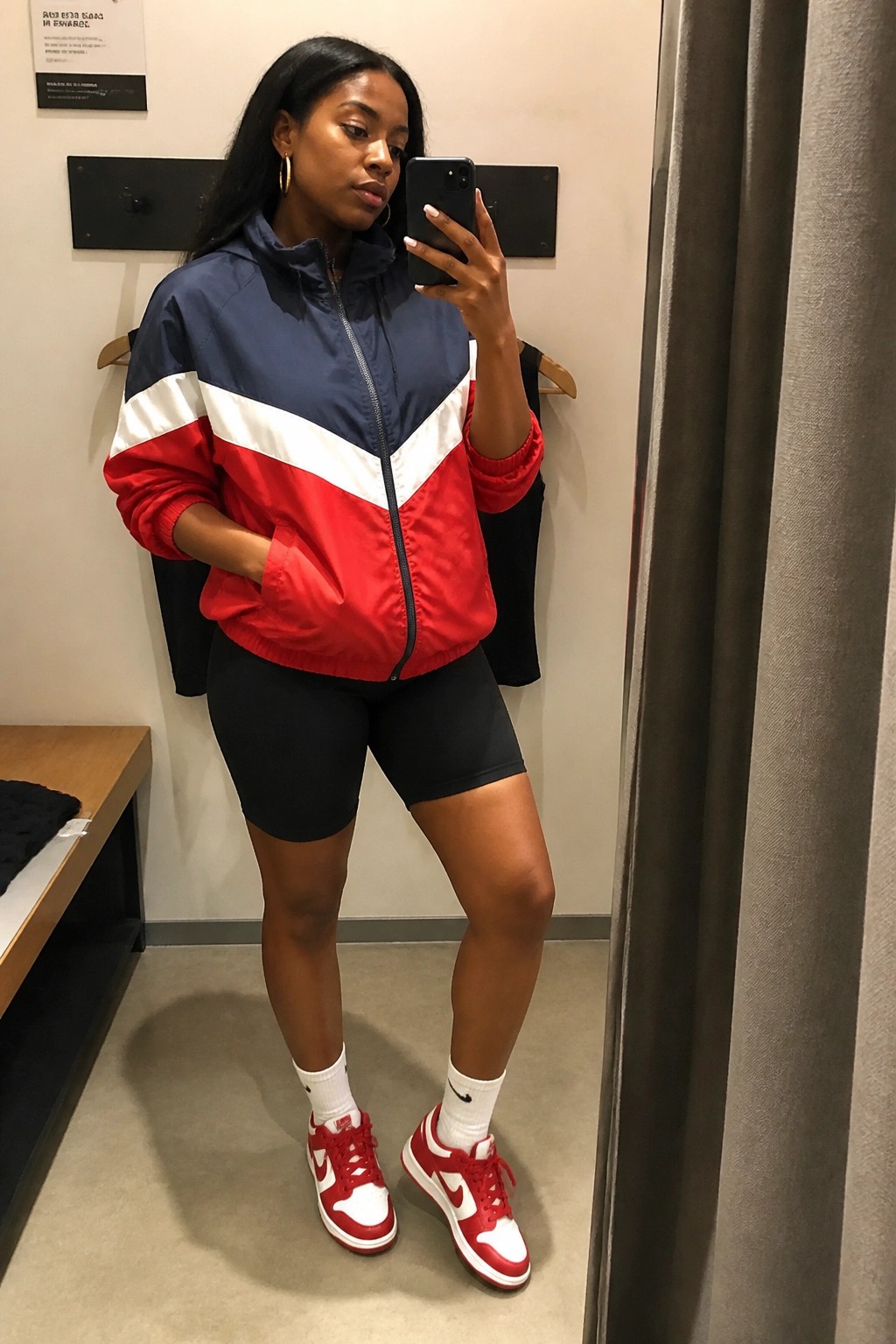Woman in navy red and white color blocked windbreaker jacket black bike shorts red and white Nike Dunk sneakers white crew socks taking mirror selfie in fitting room.