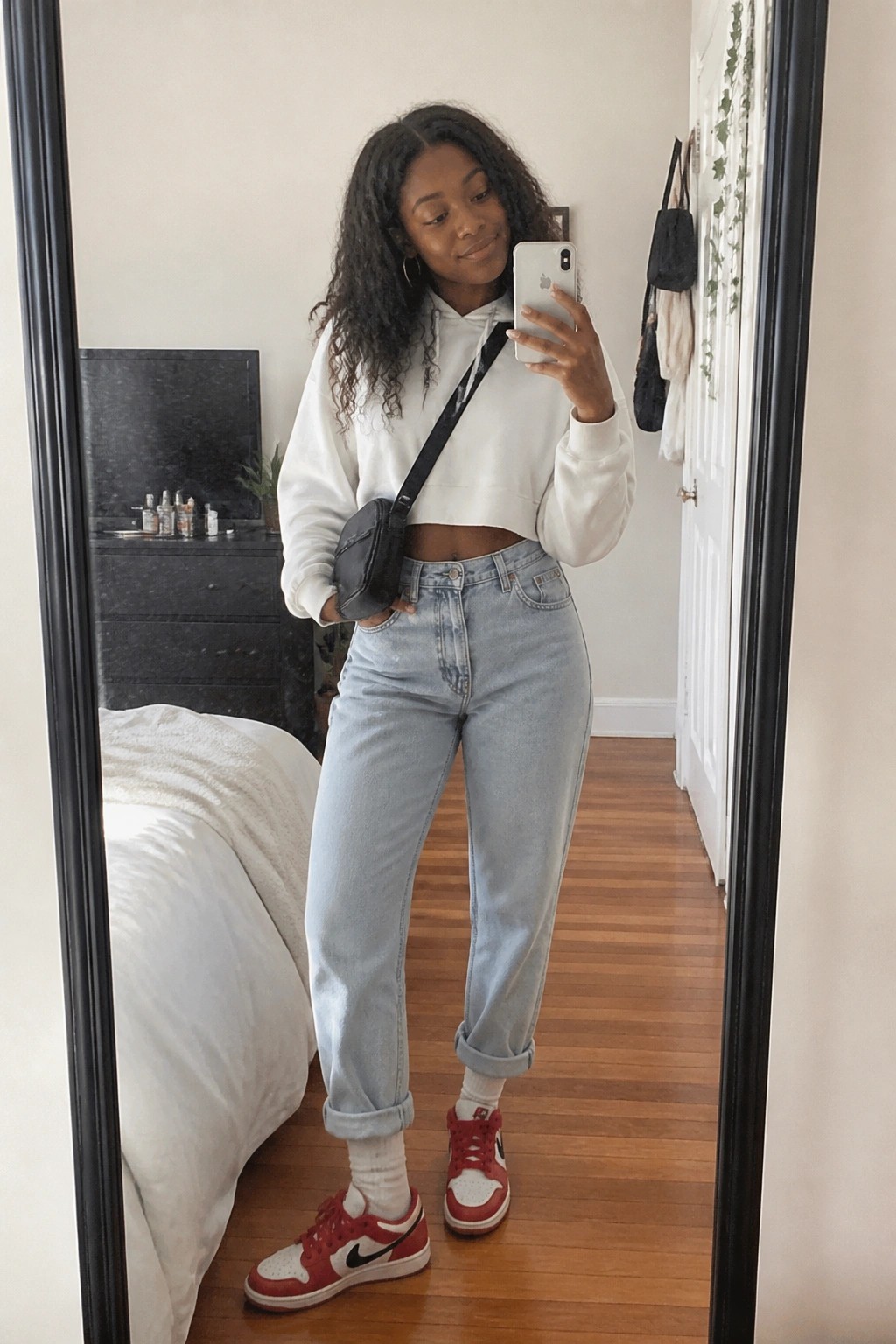 Woman with curly hair in white cropped hoodie and cuffed jeans taking mirror selfie