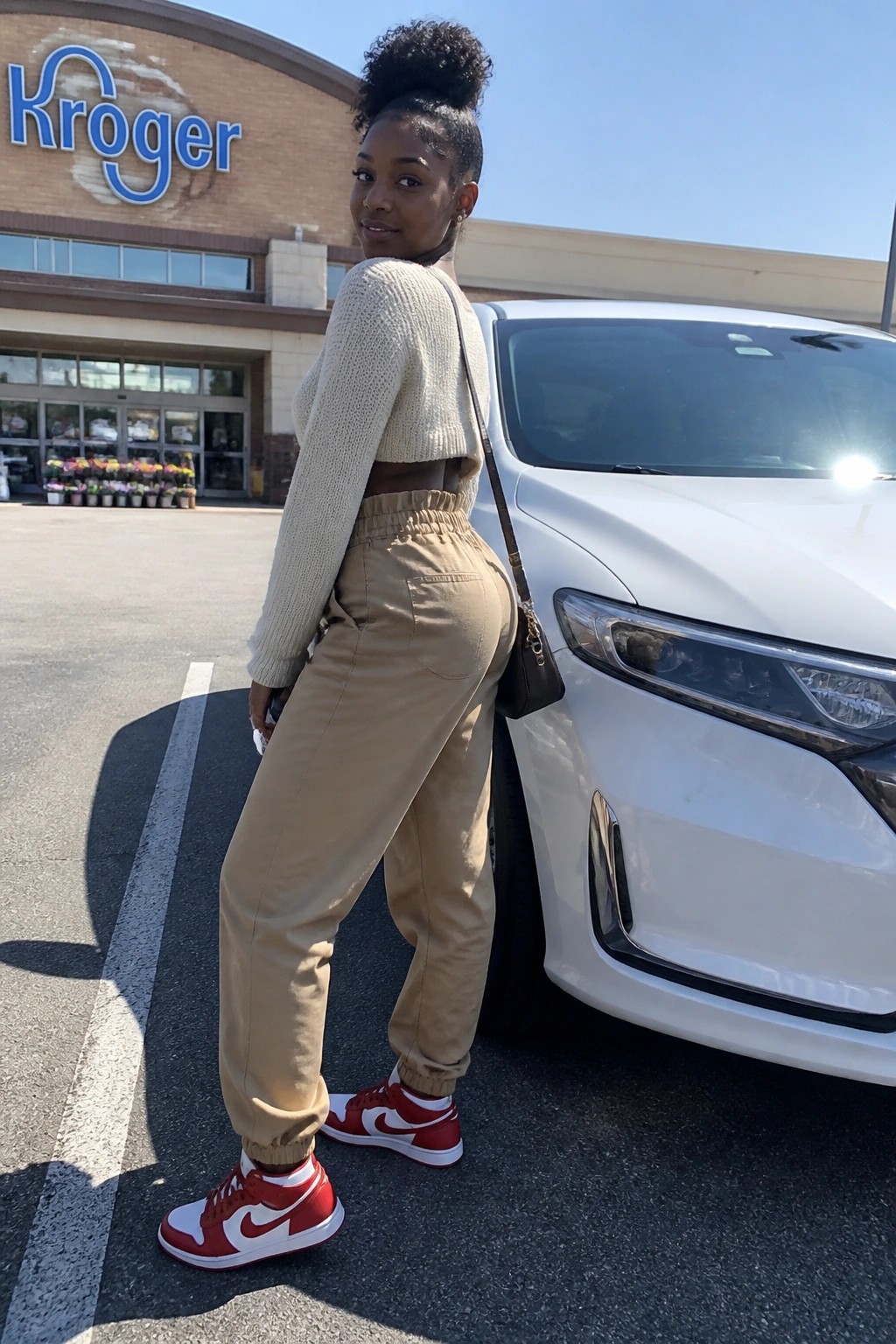 Woman in beige crop sweater and tan joggers leaning on white car outside Kroger