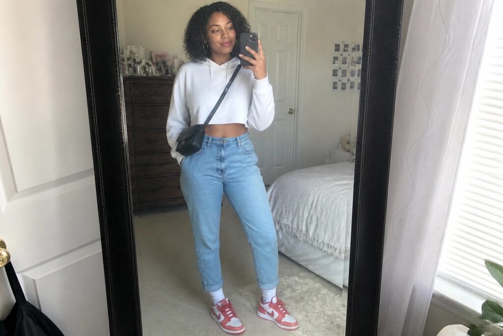 Woman with curly hair in white cropped hoodie and cuffed jeans taking mirror selfie