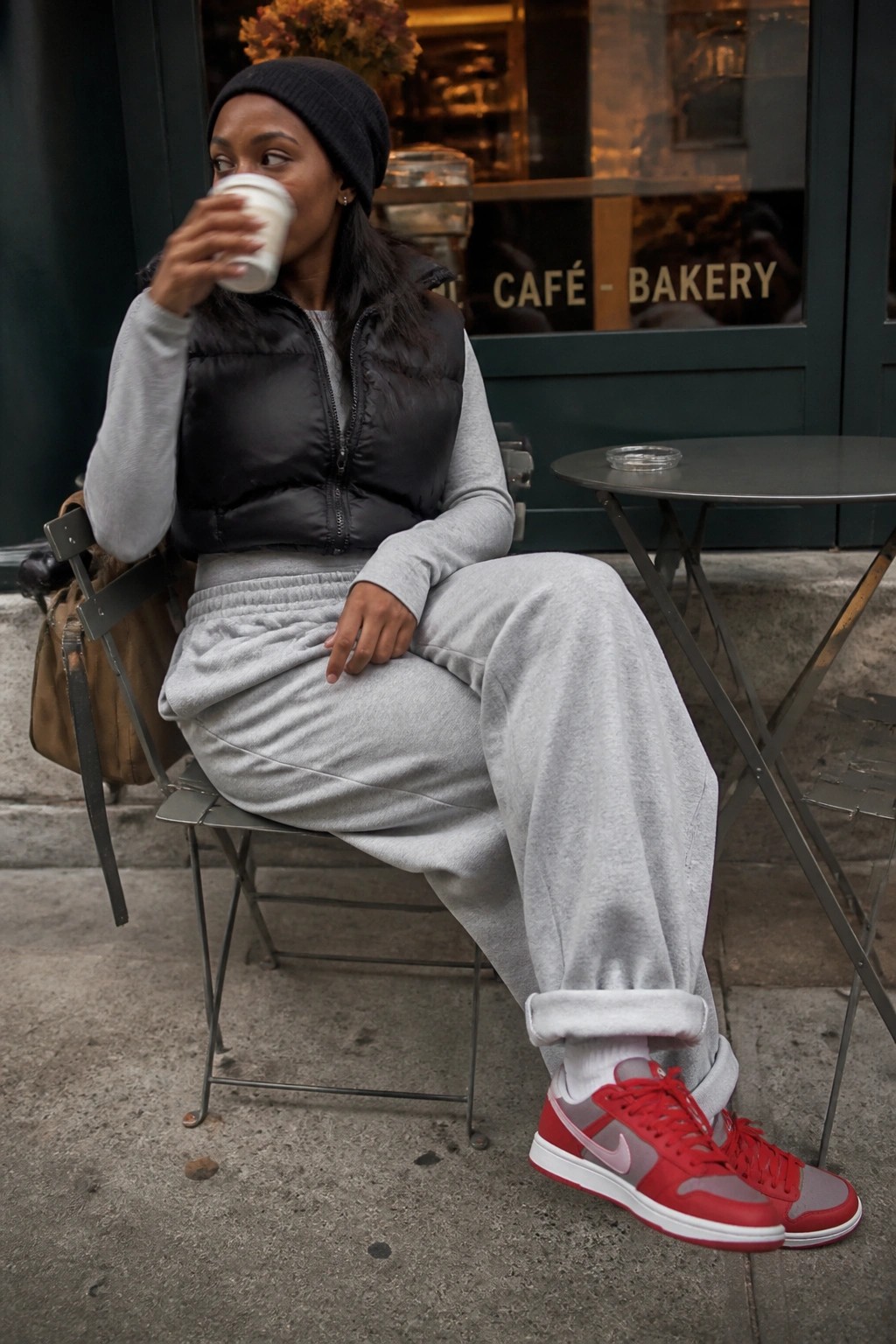 Woman in grey sweatsuit and black vest sips coffee outside café.