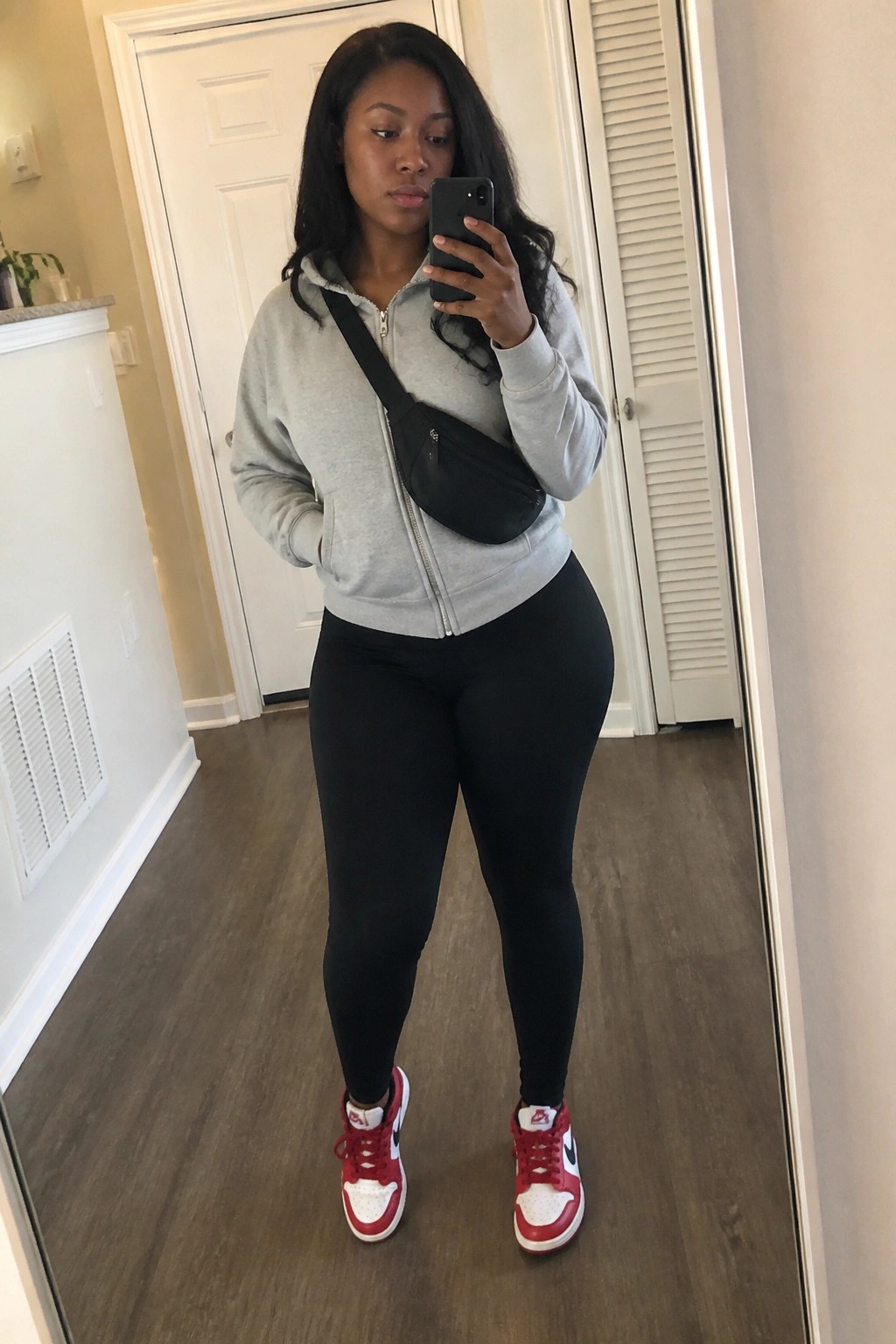 Woman in gray zip-up hoodie, black leggings, red sneakers taking mirror selfie