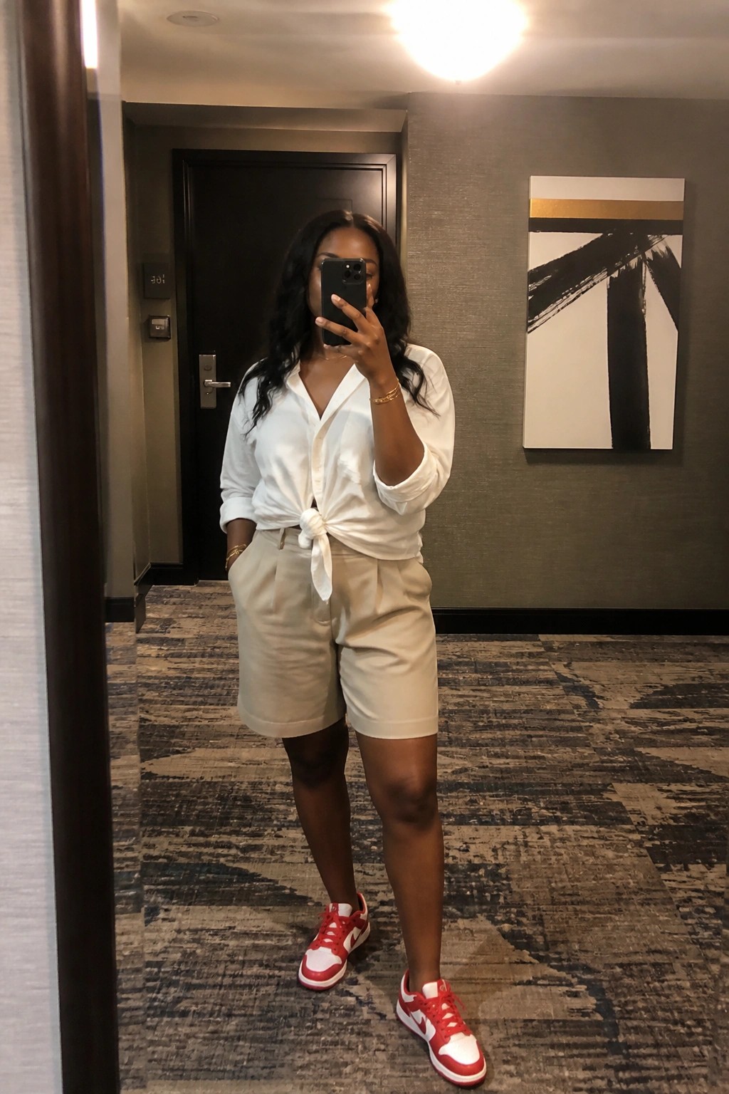 Woman in white tied shirt and beige shorts takes mirror selfie wearing red sneakers in hallway