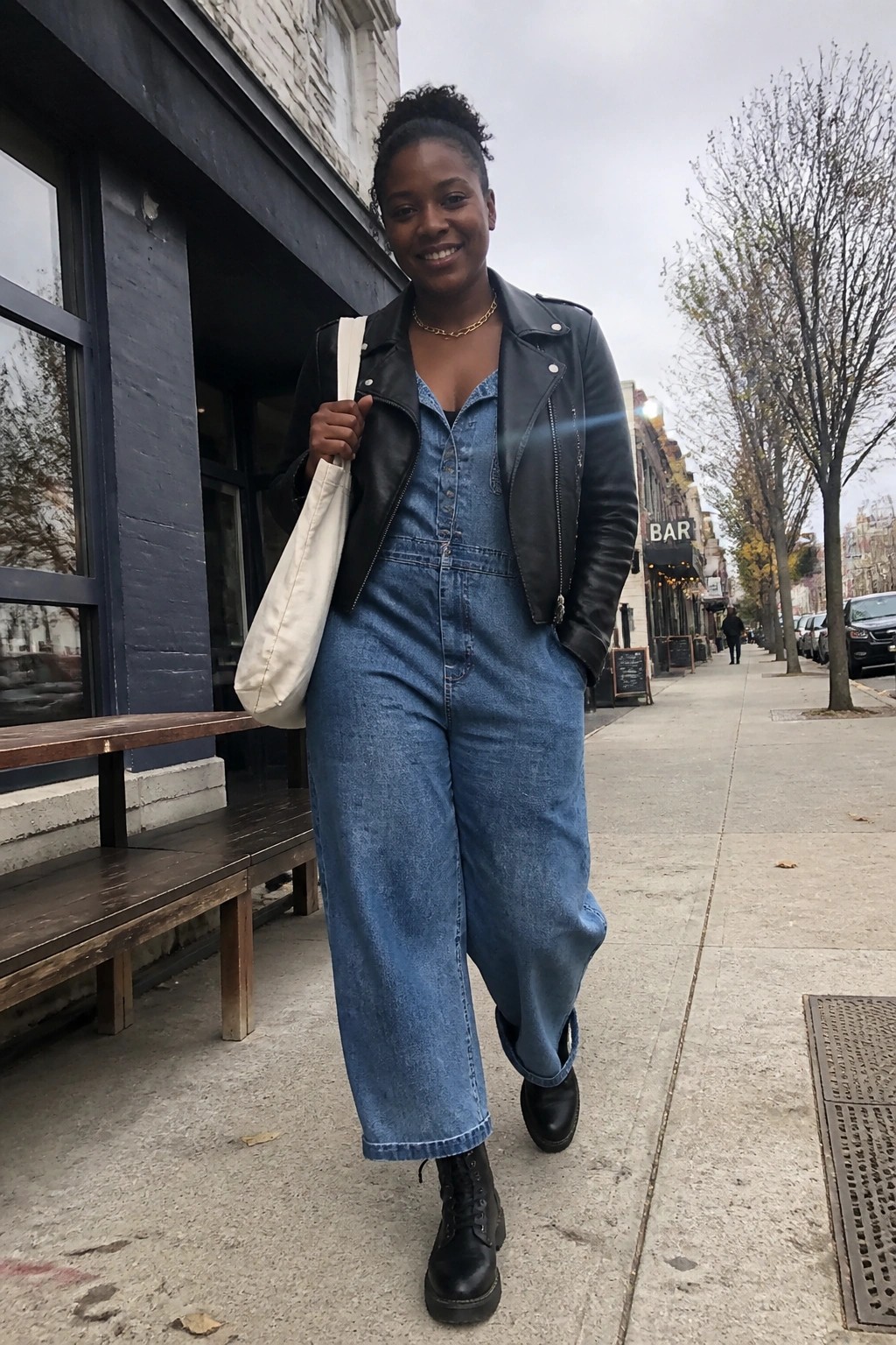Woman in blue denim jumpsuit and black leather jacket walking city sidewalk with tote bag