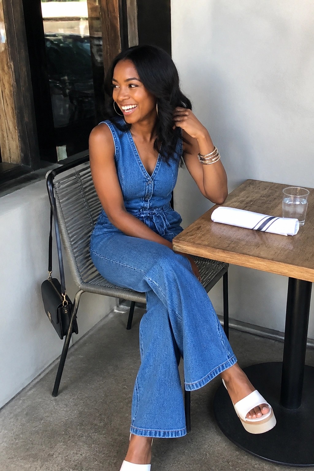 Smiling woman in blue denim jumpsuit seated at outdoor wooden cafe table