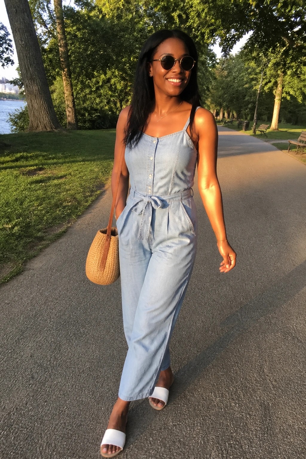 Smiling woman in light blue denim jumpsuit with woven bag on sunny park path