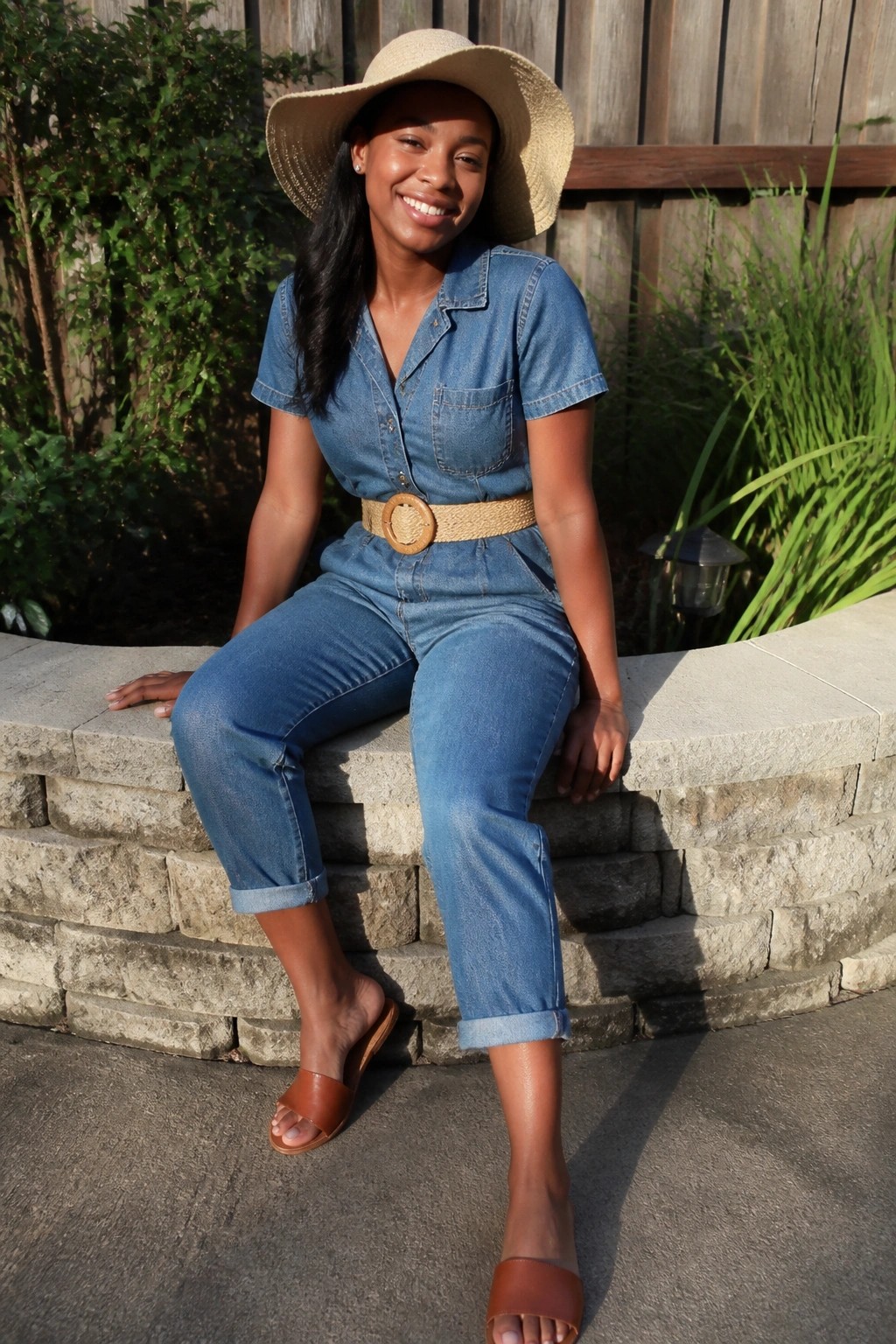 Black woman in short sleeve blue denim jumpsuit with woven belt wooden buckle cuffed ankles brown slides and straw hat seated on stone steps