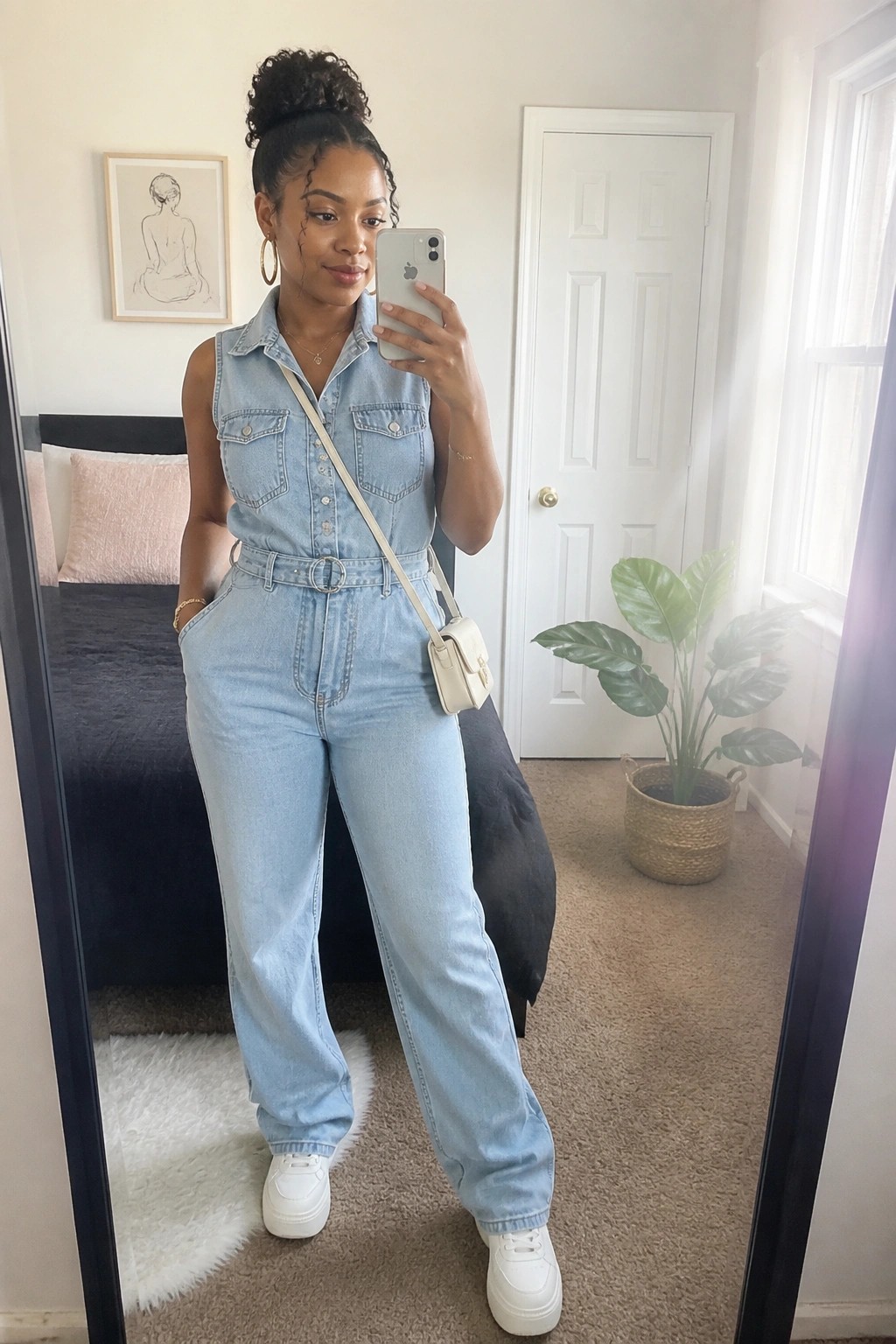 Woman in light blue denim jumpsuit taking mirror selfie in bedroom