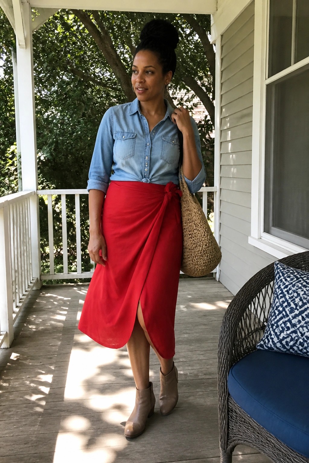 Woman in blue denim shirt and red wrap skirt with straw bag on porch