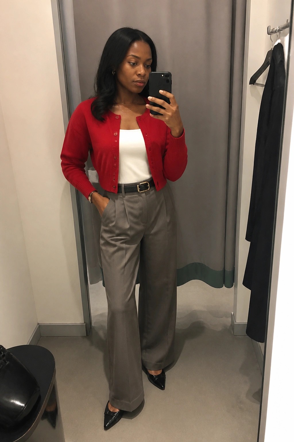 Woman in red cardigan and gray trousers taking mirror selfie in fitting room