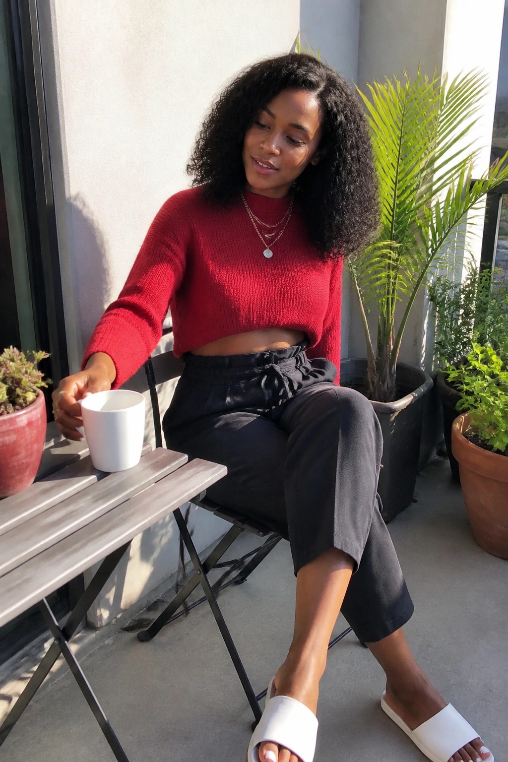 Curly-haired woman in red sweater and black pants holding white mug on sunny balcony.