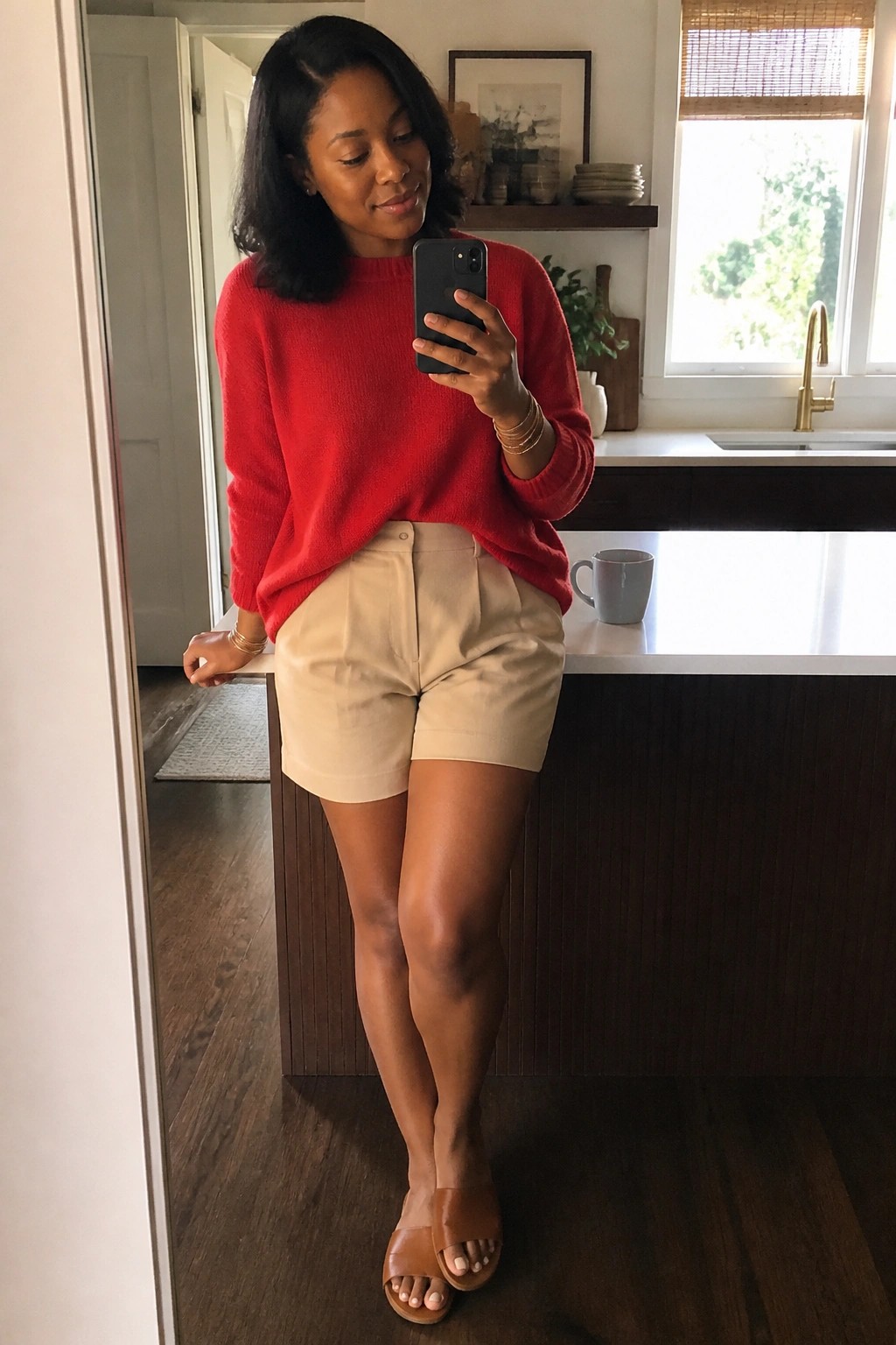 Woman in red sweater and beige shorts taking mirror selfie in kitchen