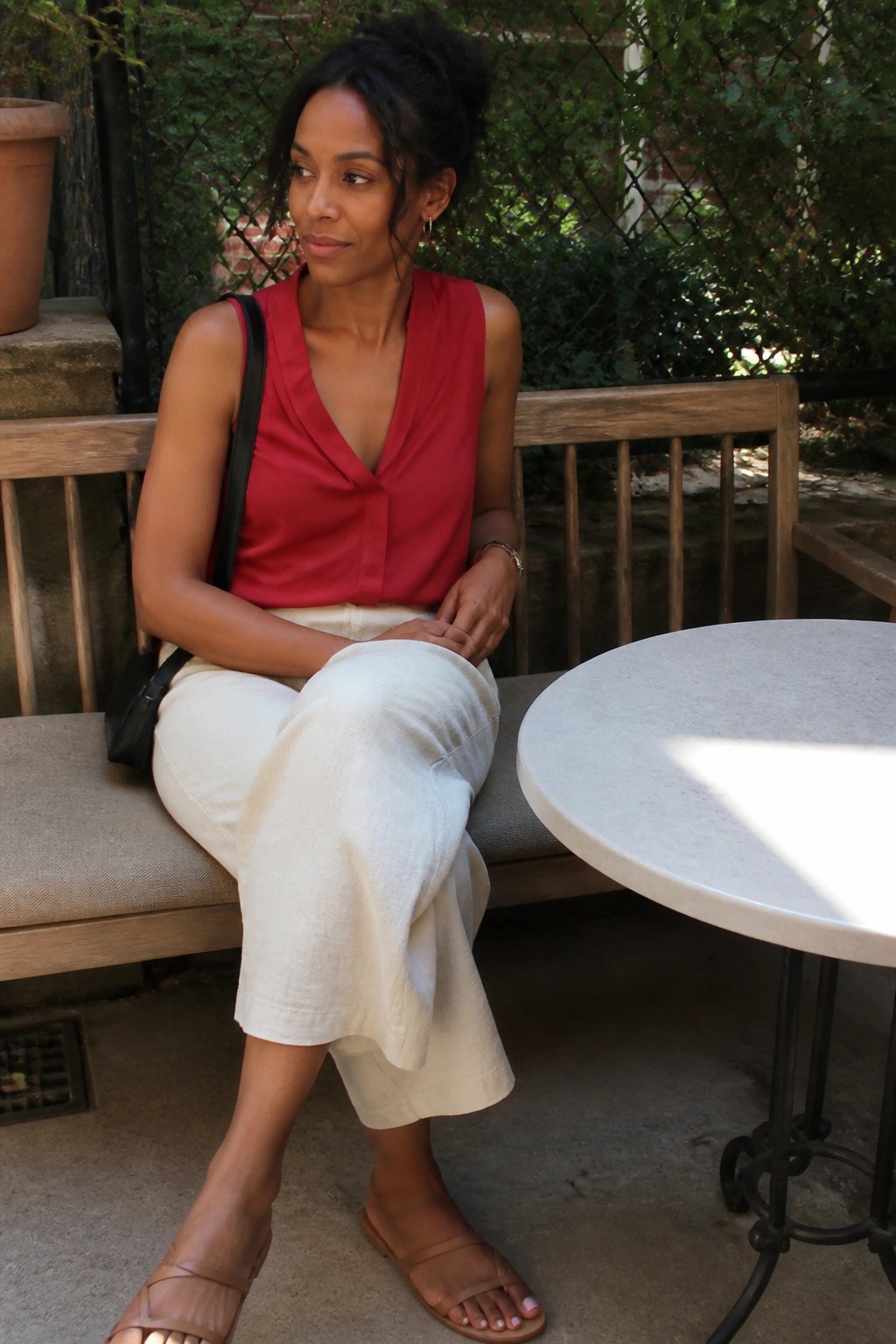 Woman in red top and cream pants seated on bench by round table outdoors