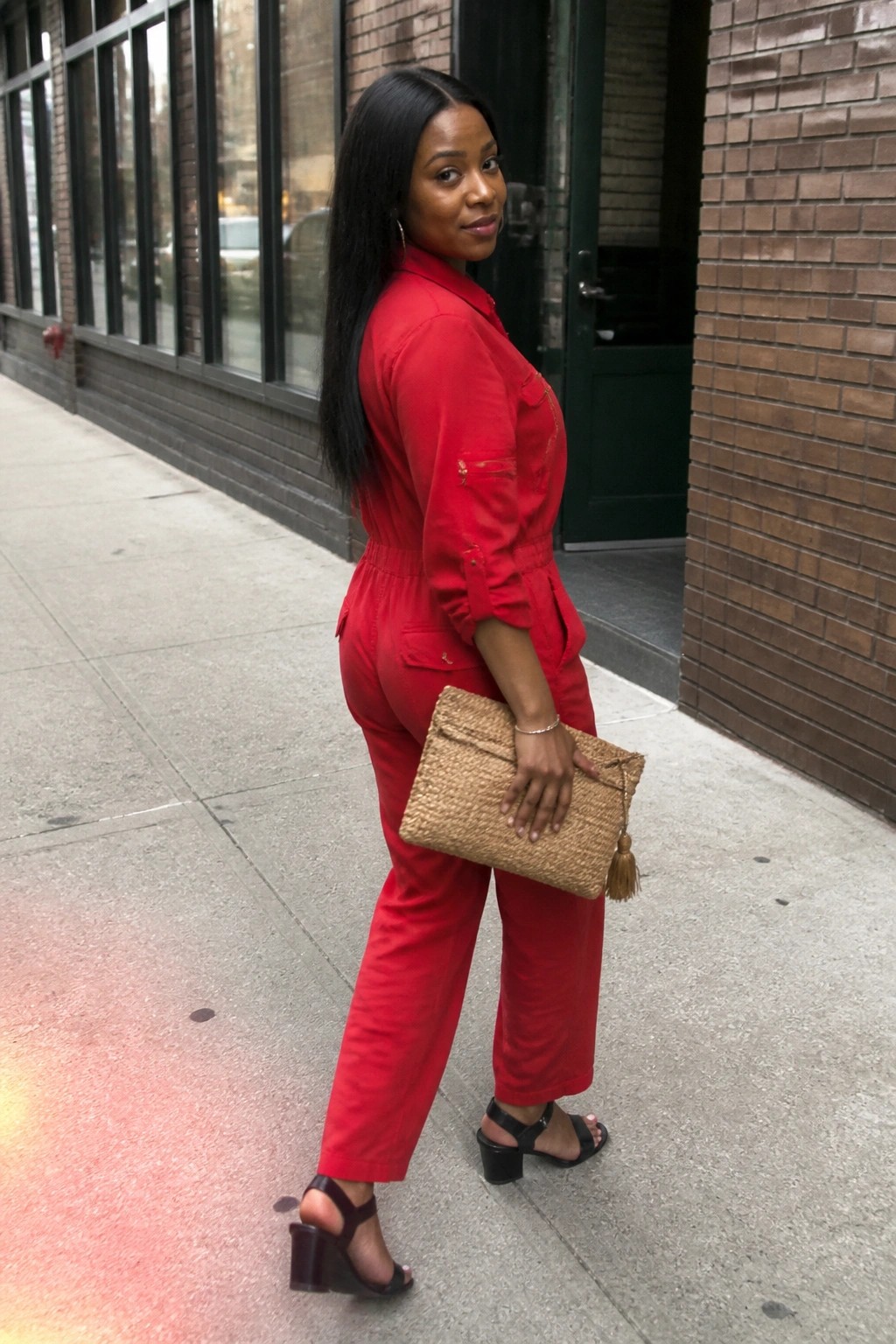 Woman in red jumpsuit holding woven clutch on urban sidewalk, glancing back