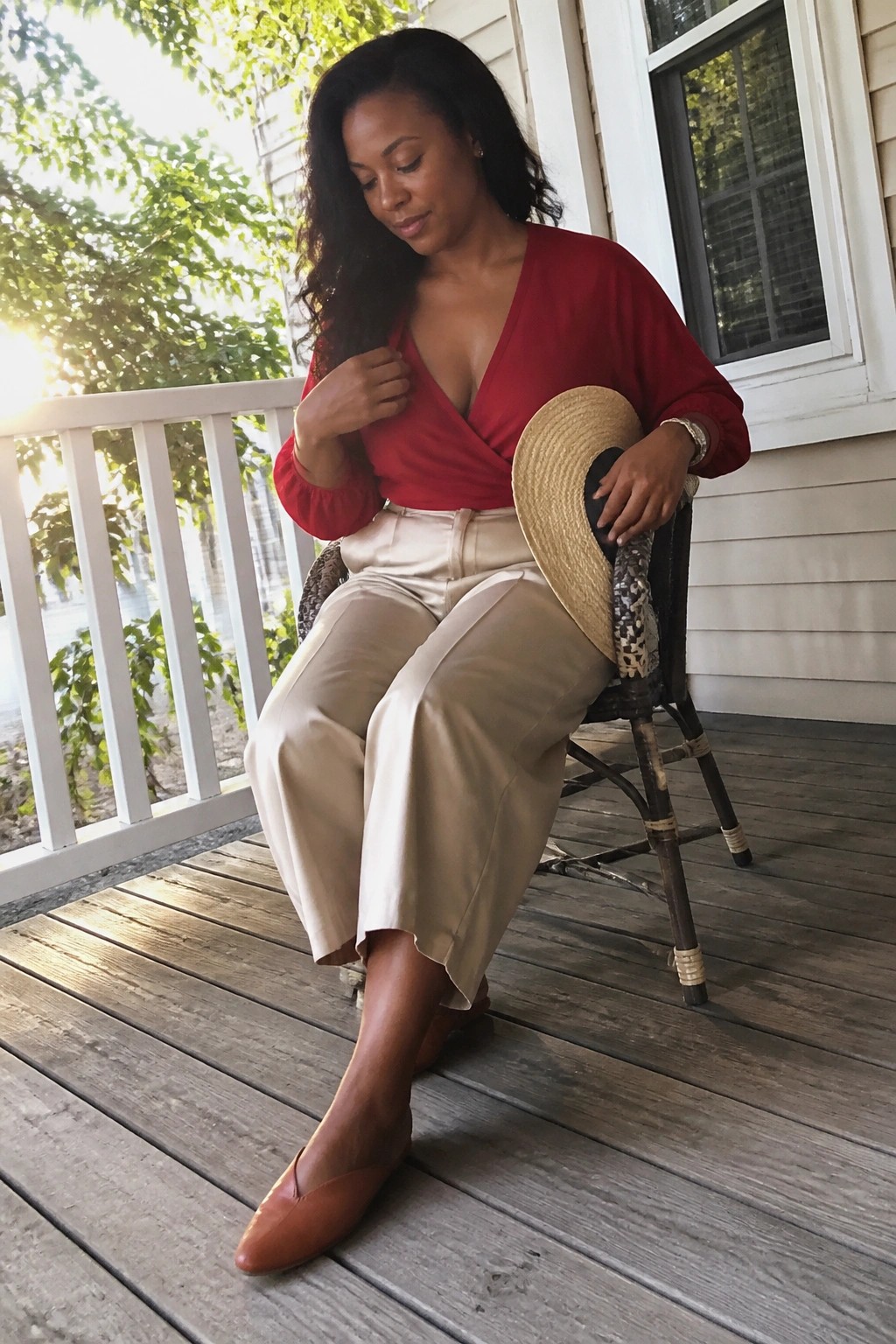 Woman in red wrap top and beige pants on porch holding straw hat