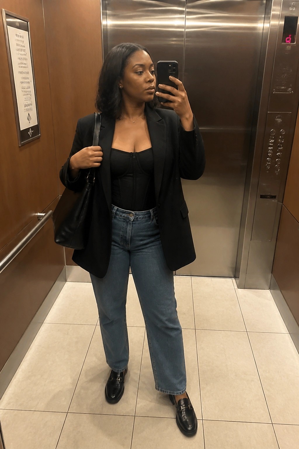 Woman in black blazer and corset taking mirror selfie in elevator with handbag