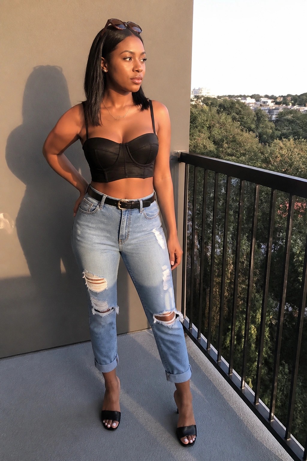 Woman in black bustier and ripped jeans posing on sunny balcony with city view.