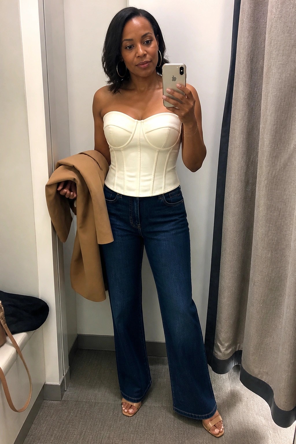 Woman in white corset top and blue wide-leg jeans taking mirror selfie holding tan jacket
