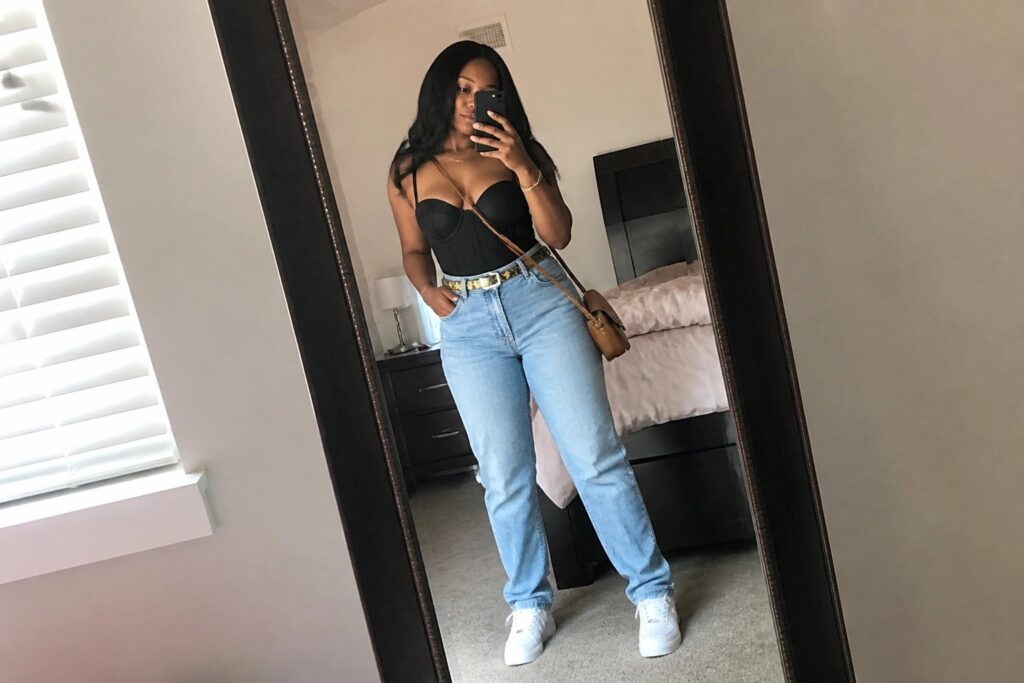 Woman in black corset top and light blue jeans taking bedroom mirror selfie