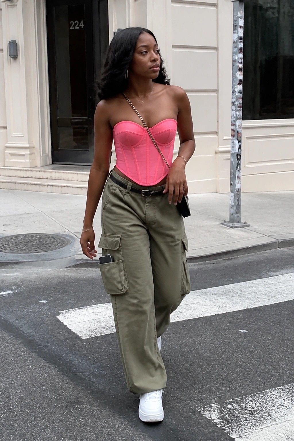 Woman in bright pink strapless corset top tucked into olive cargo pants with black belt, white sneakers, and gold chain strap bag on urban sidewalk.