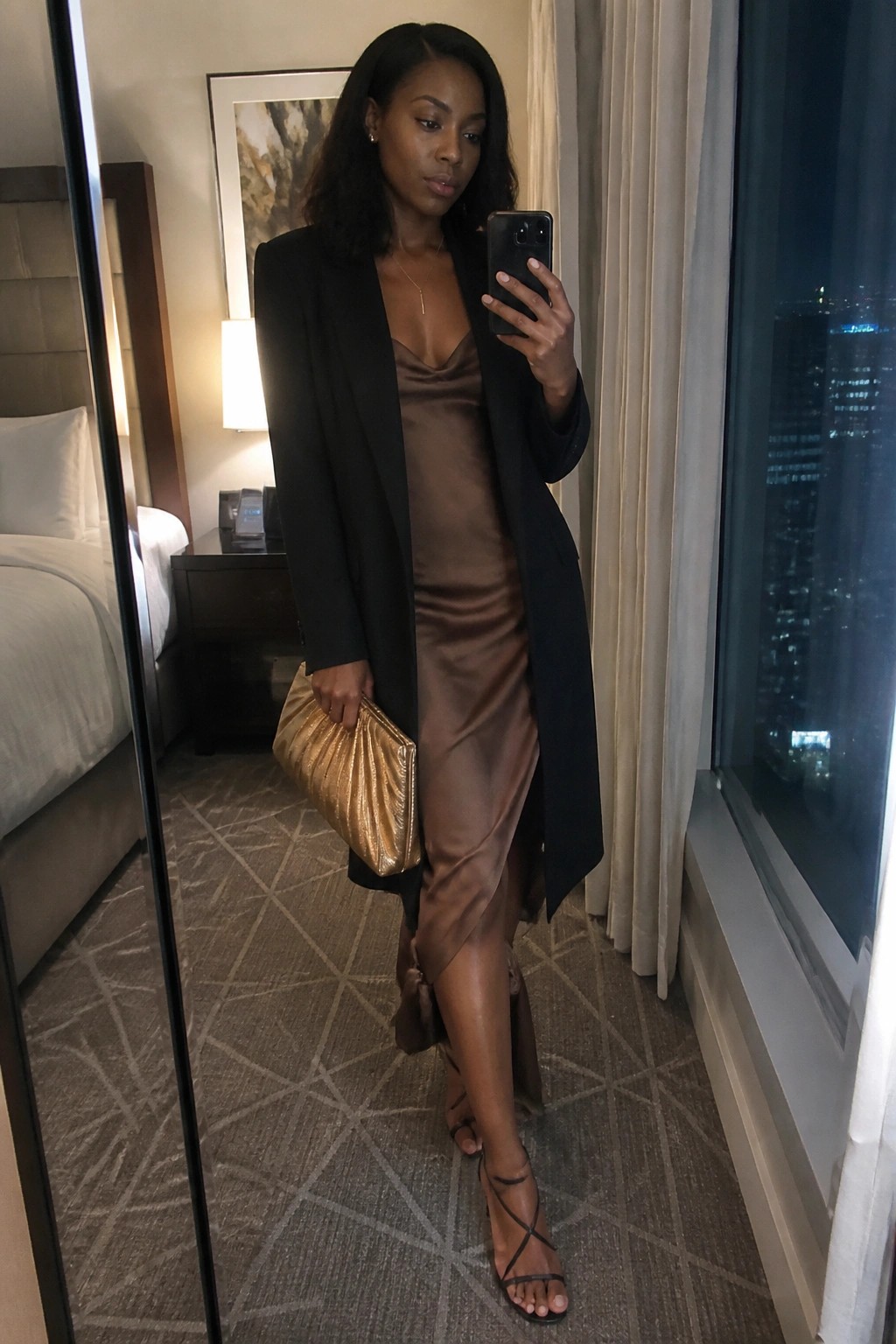 Woman in black blazer over brown satin dress taking mirror selfie in hotel room