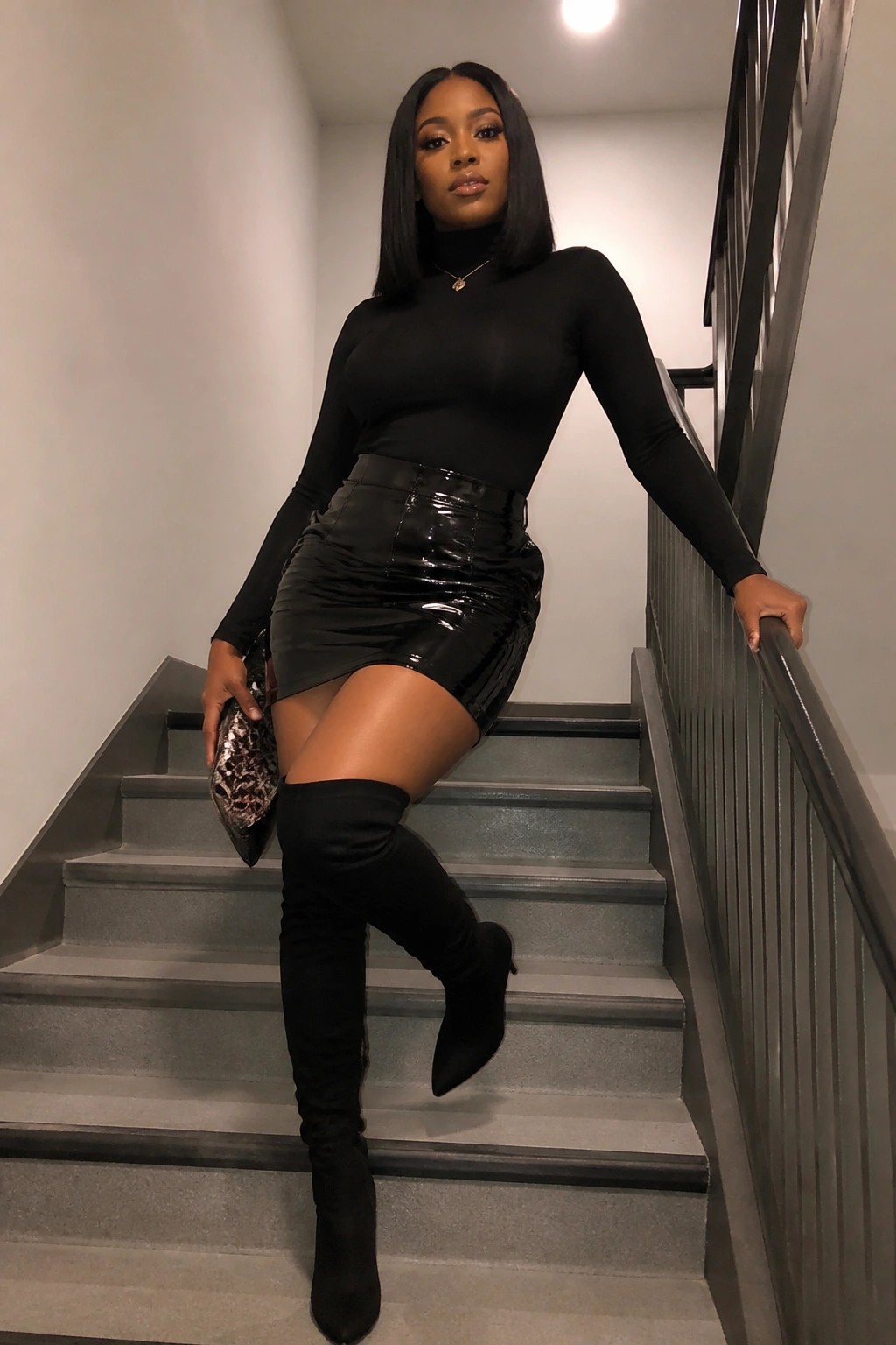 Woman in black turtleneck and glossy mini skirt posing on gray staircase in thigh-high boots