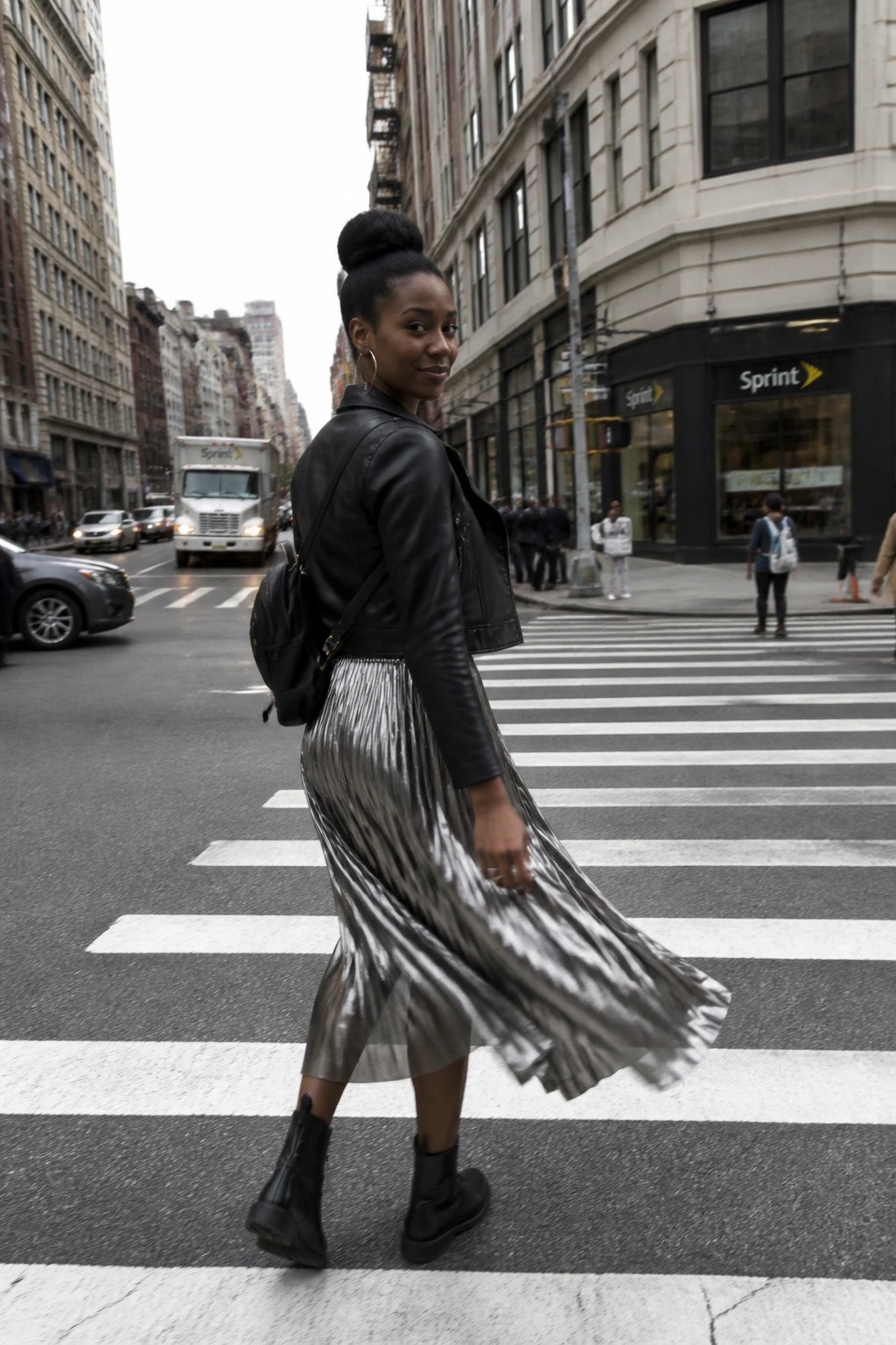 Woman in black leather jacket and pleated silver skirt crossing city crosswalk, looking back.
