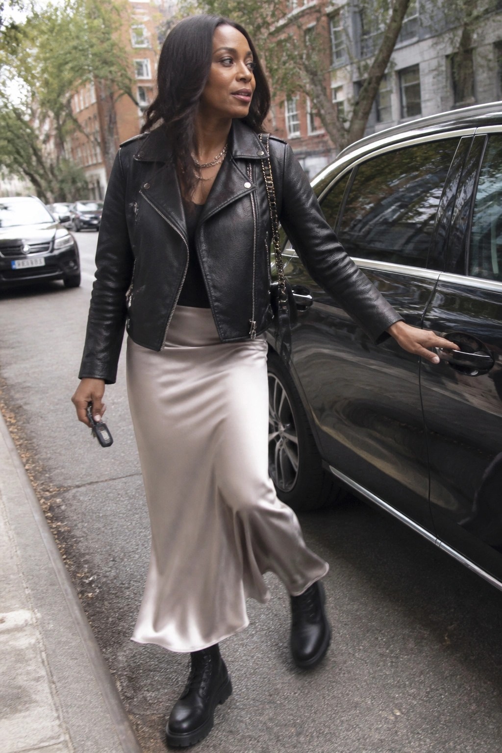 Woman in black leather jacket and beige satin skirt by black car on street