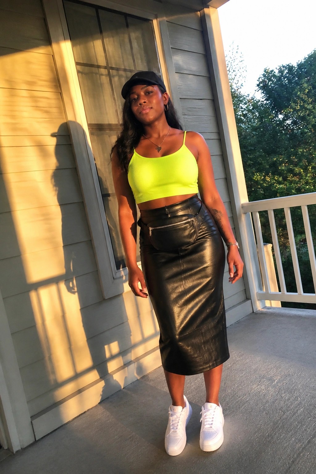 Woman in neon yellow crop top and black leather skirt on sunny wooden porch.