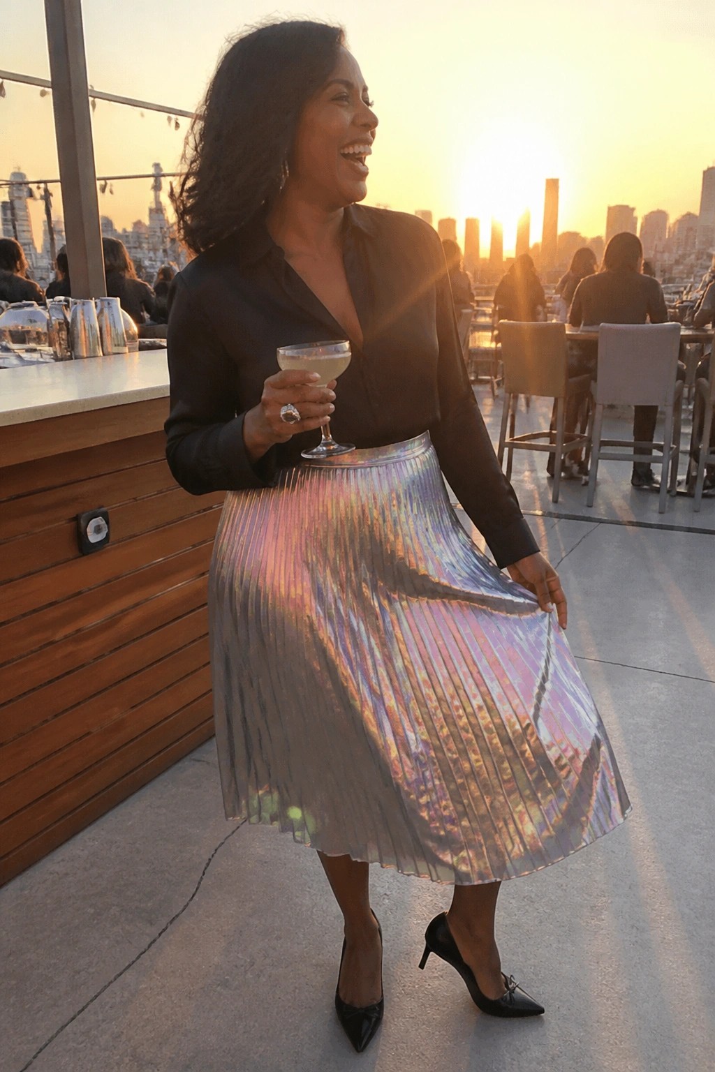 Woman in black blouse and iridescent pleated skirt holding cocktail on sunset rooftop