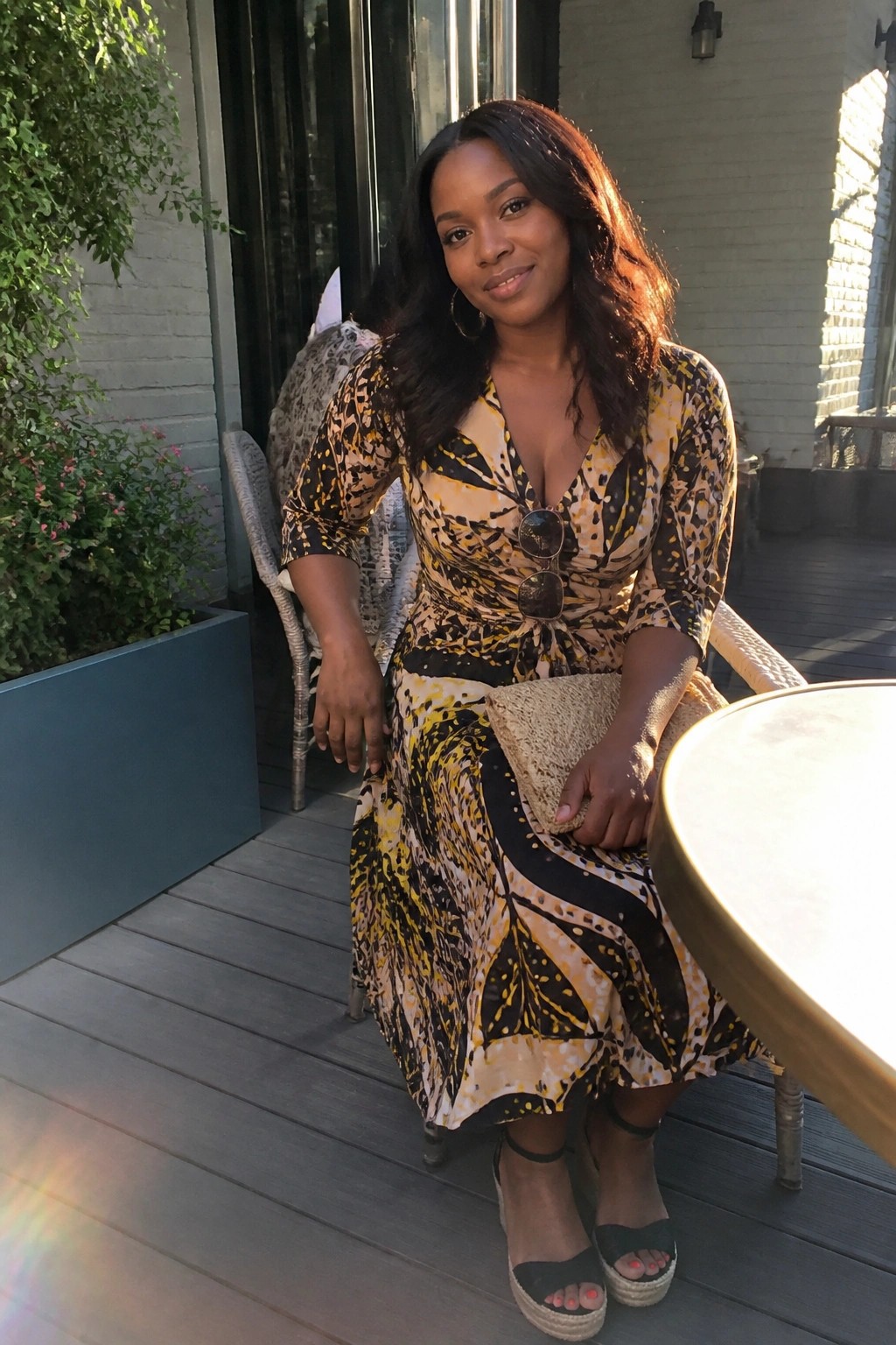 Woman in a yellow black and beige abstract print midi wrap dress with three quarter sleeves cinched waist and V neck paired with black wedge espadrilles and a woven clutch held in one hand.