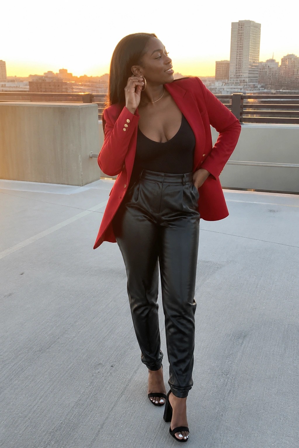 Woman in red blazer and black leather pants posing on city rooftop at sunset