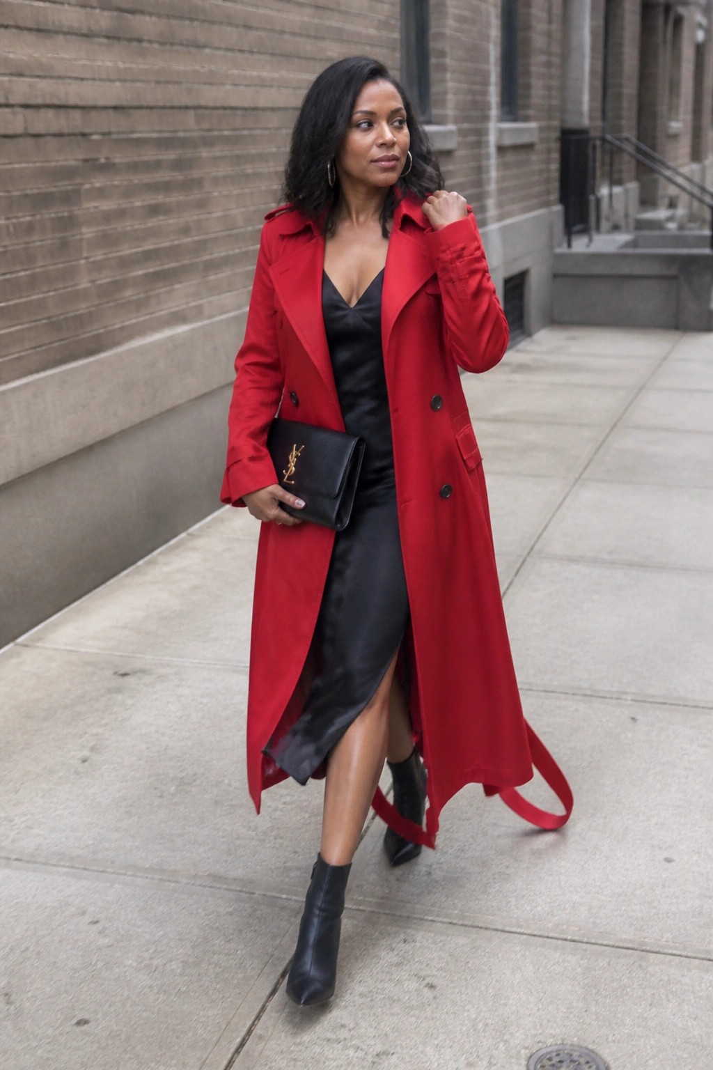 Woman in red trench coat over black dress holding YSL clutch on sidewalk.