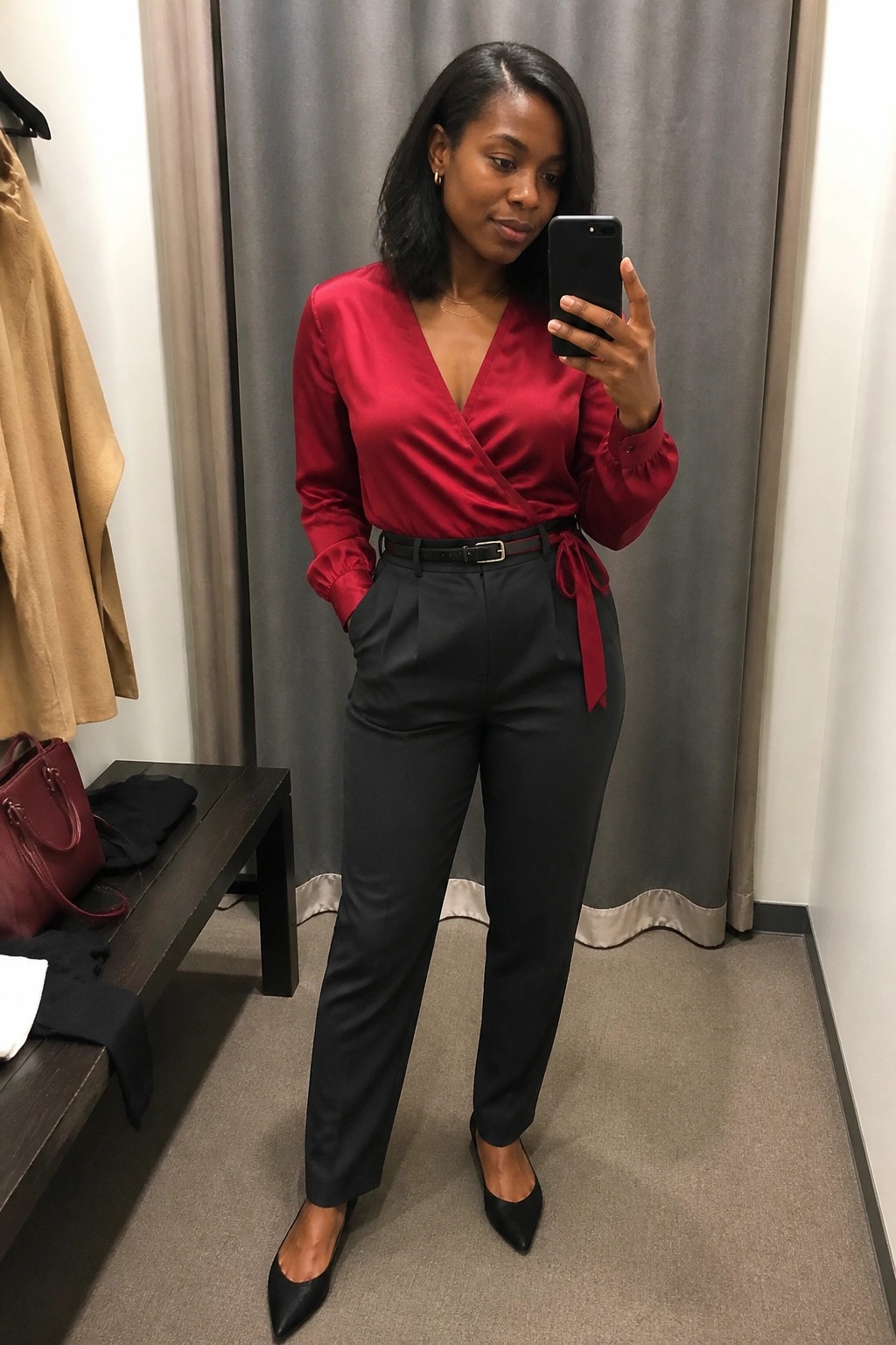 A woman in a red satin wrap blouse with black pleated trousers, black belt, and pointed black flats taking a mirror selfie.
