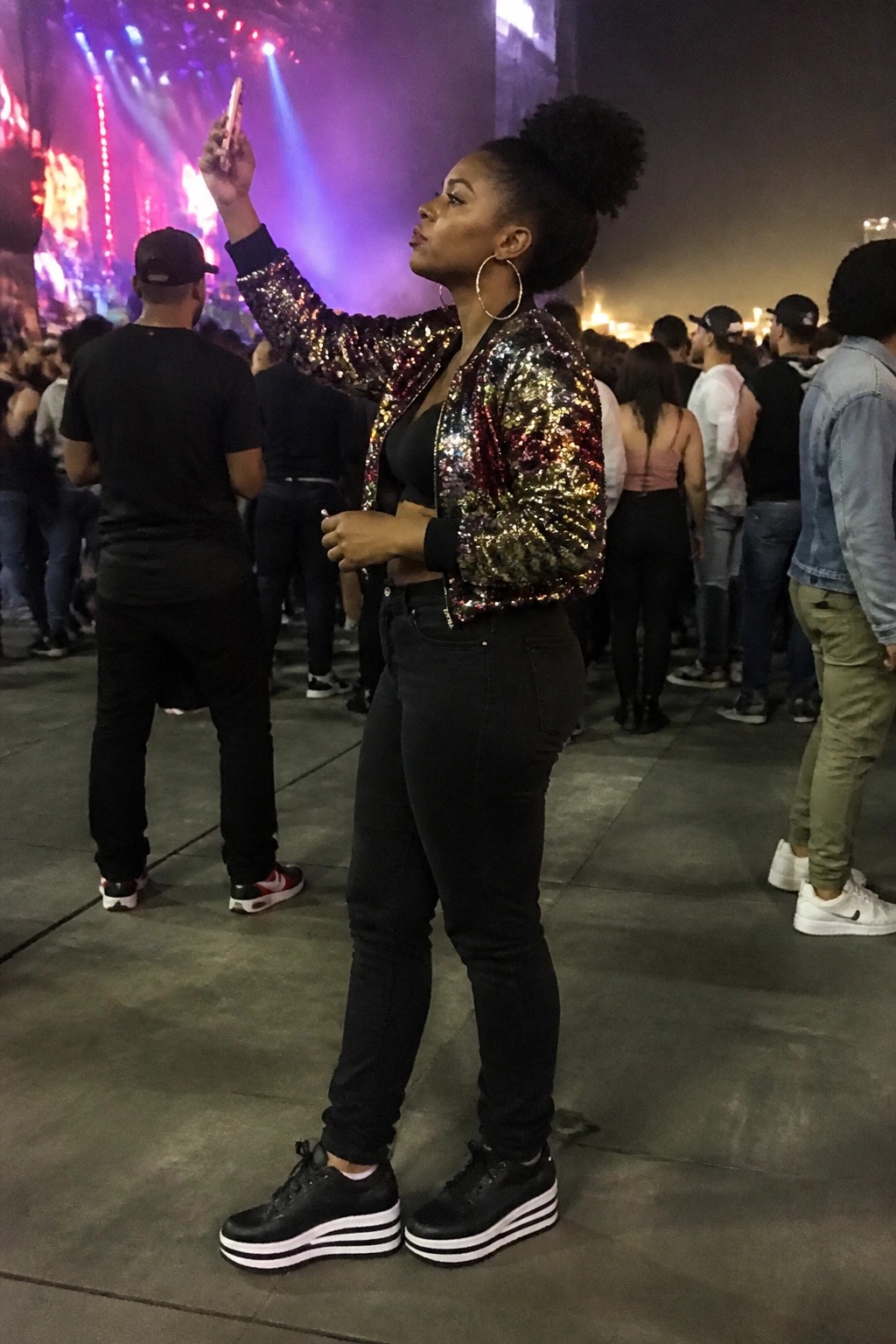 Woman in sparkly sequined jacket takes selfie at concert with crowd and stage lights