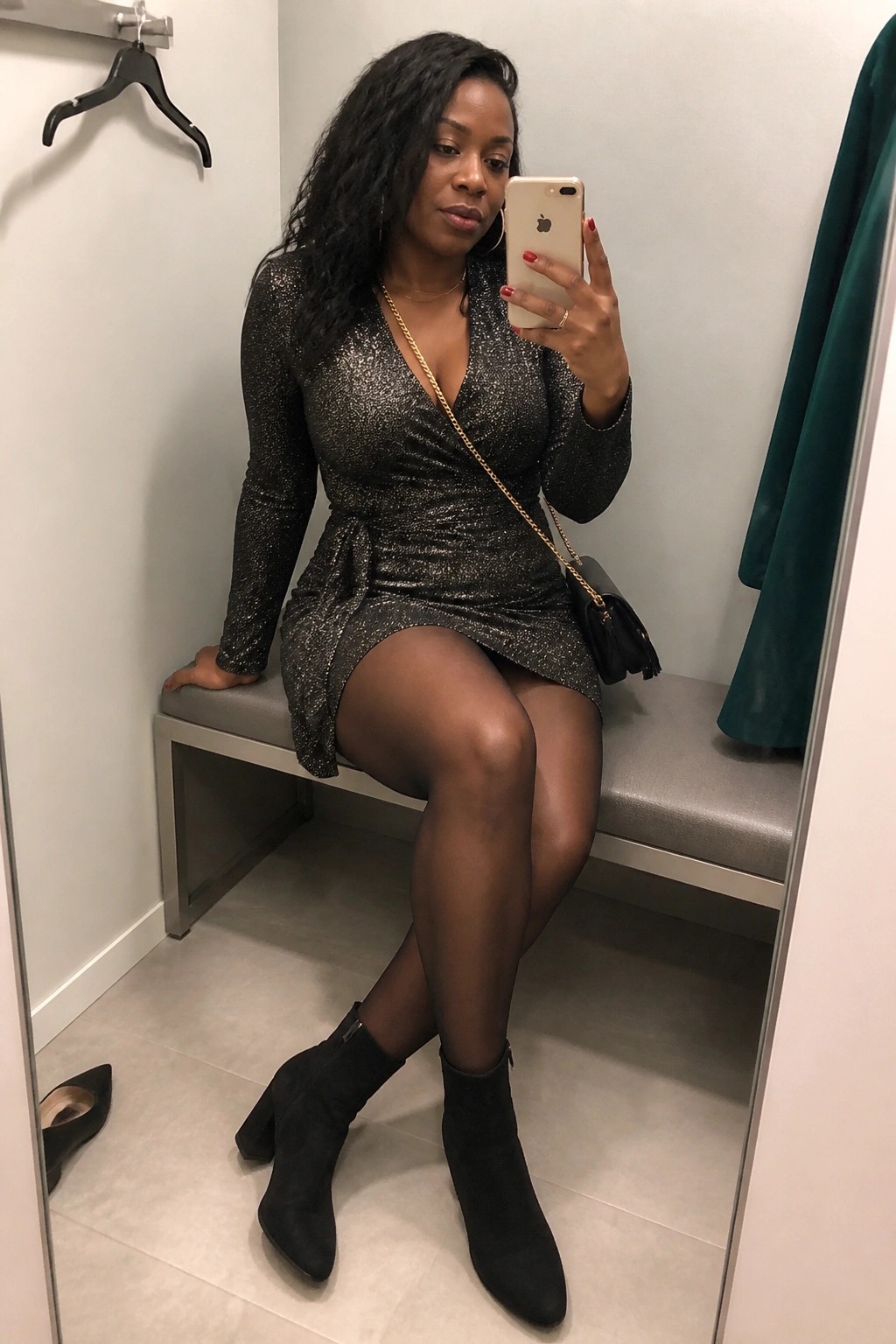 Woman wearing a short black sparkly wrap dress with long sleeves, sheer black tights, and black ankle boots while holding a phone in a dressing room.