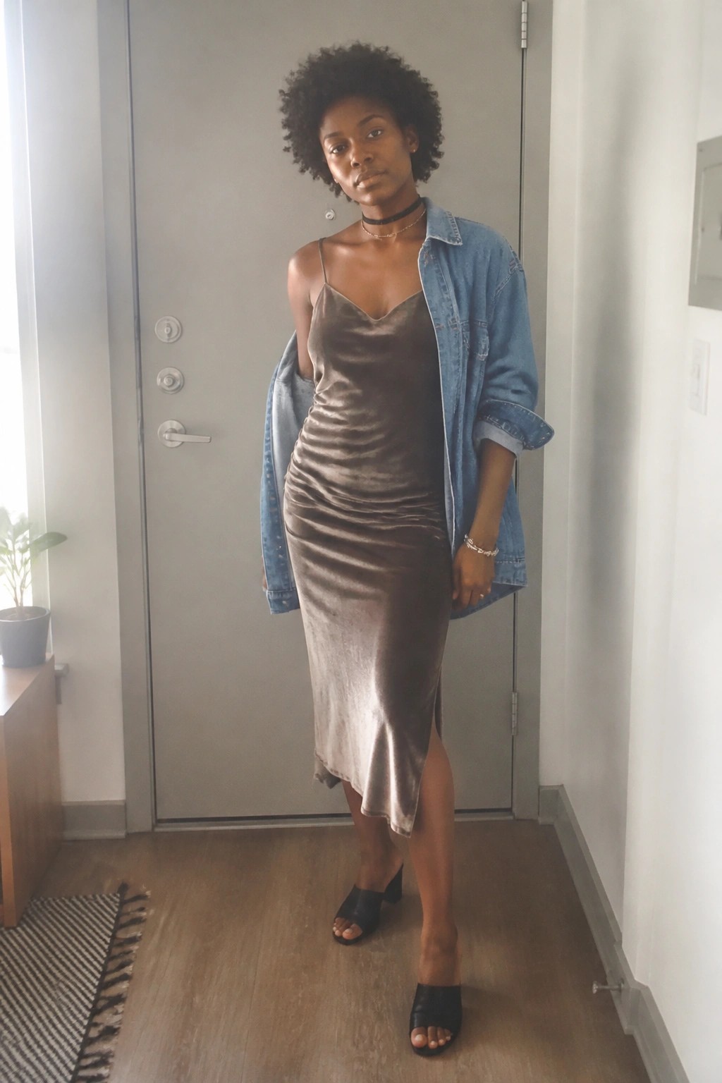 Woman in taupe velvet slip dress and blue denim jacket standing indoors.