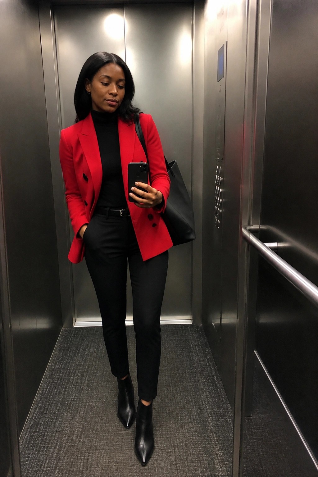 Woman in red blazer taking phone selfie inside metallic elevator with black bag