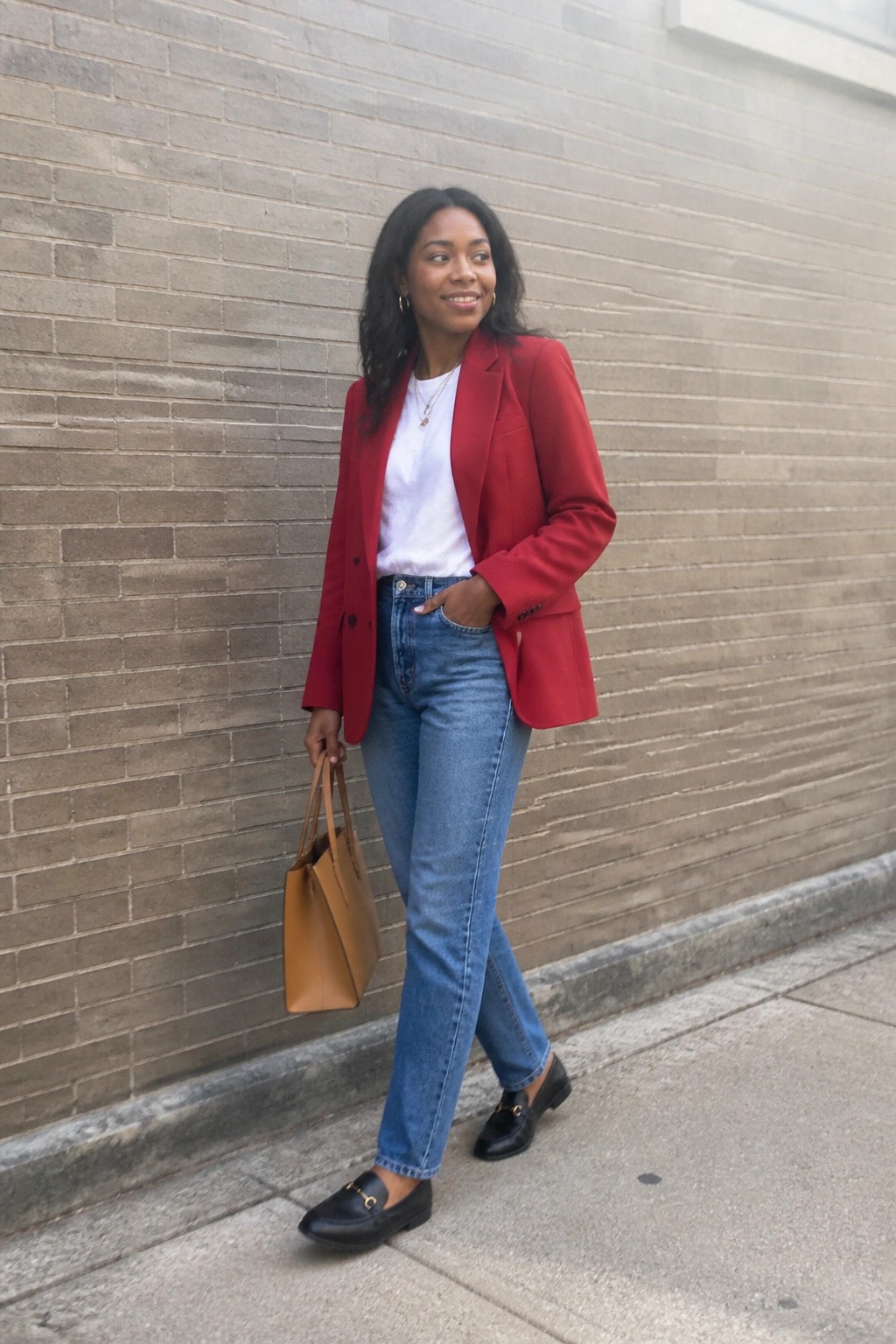 Woman in red blazer and jeans holding tan bag by brick wall