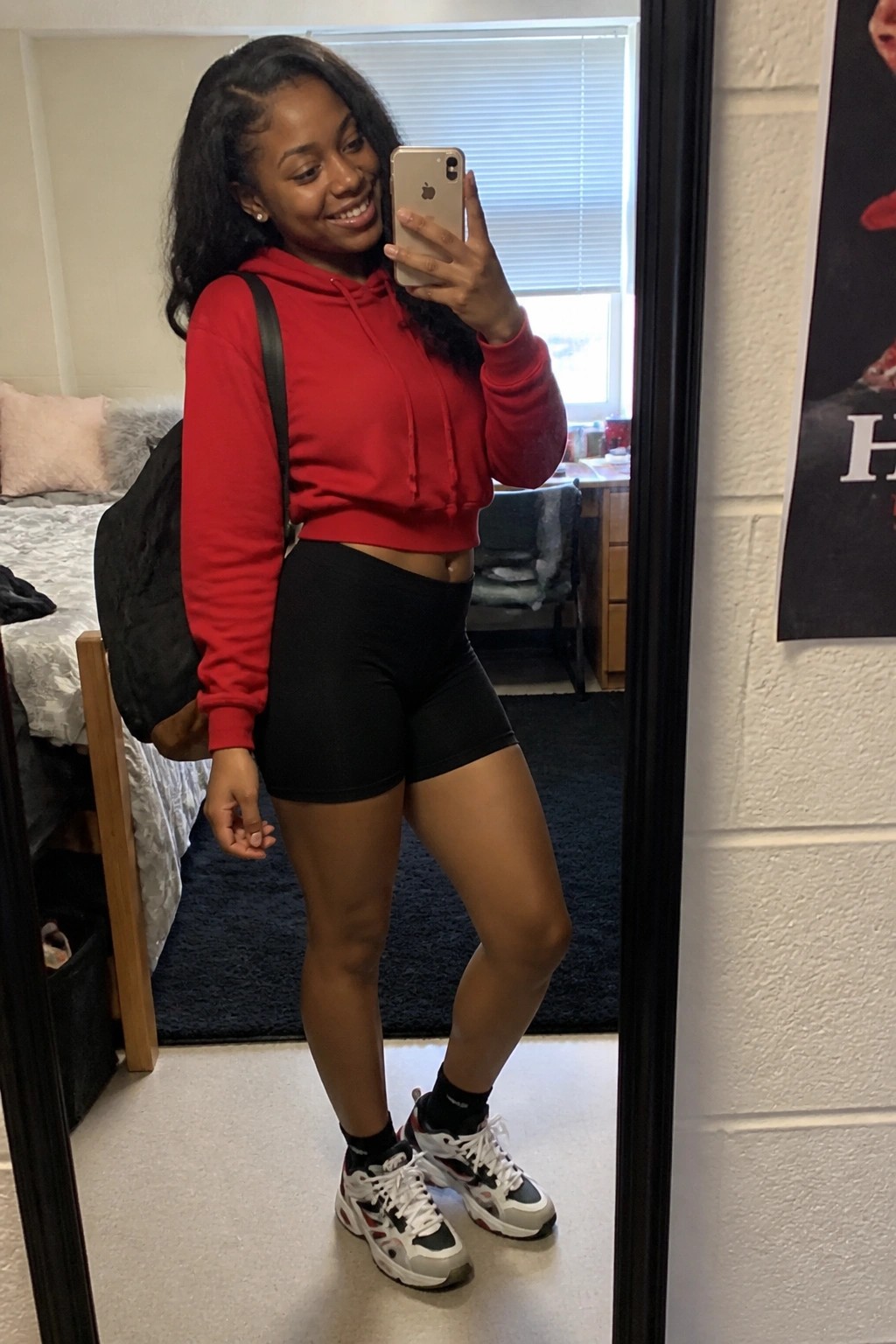 Young woman in red cropped hoodie and black shorts takes mirror selfie in dorm room