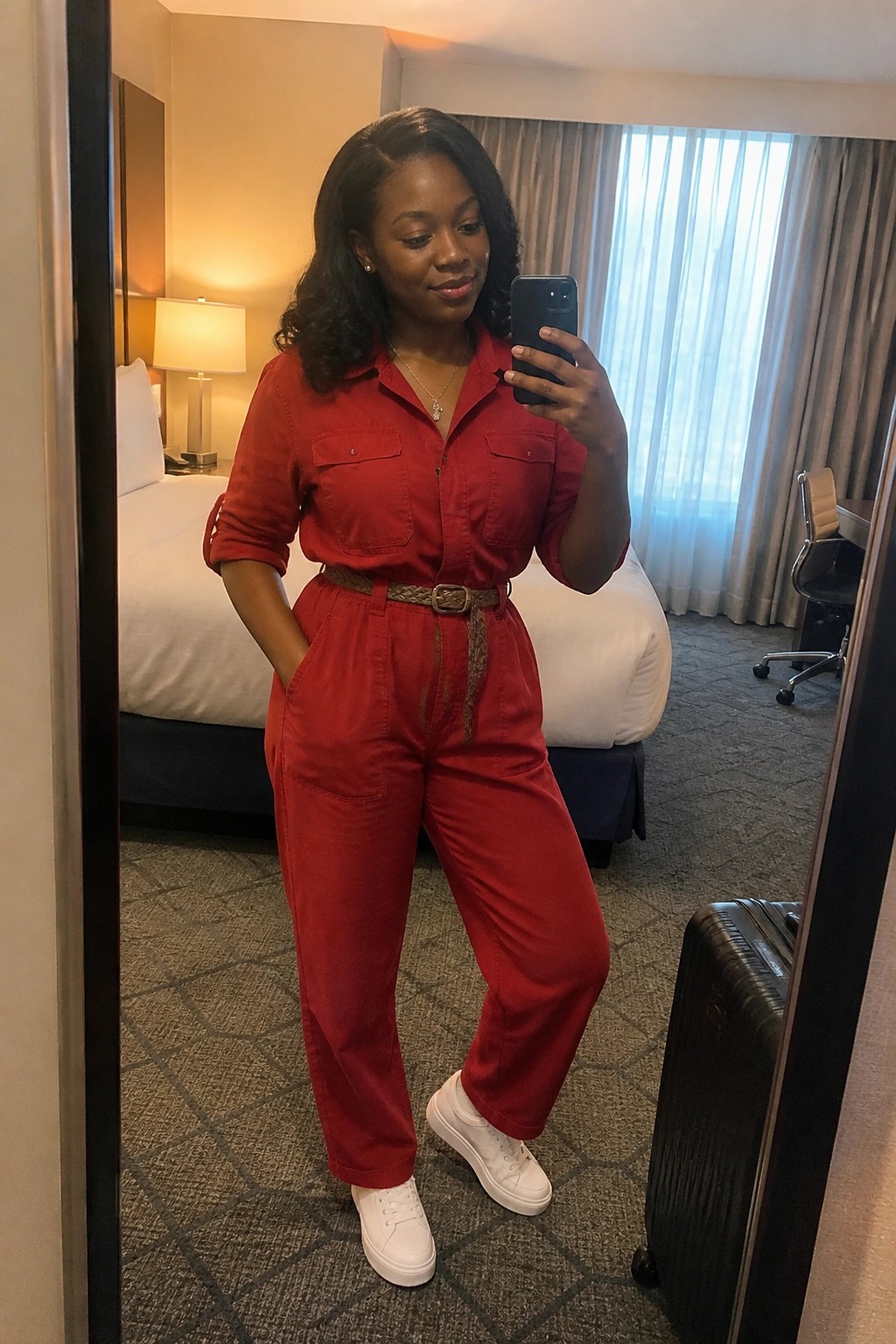 Woman wearing a red utility jumpsuit with a brown belt and white sneakers taking a mirror selfie in a hotel room.