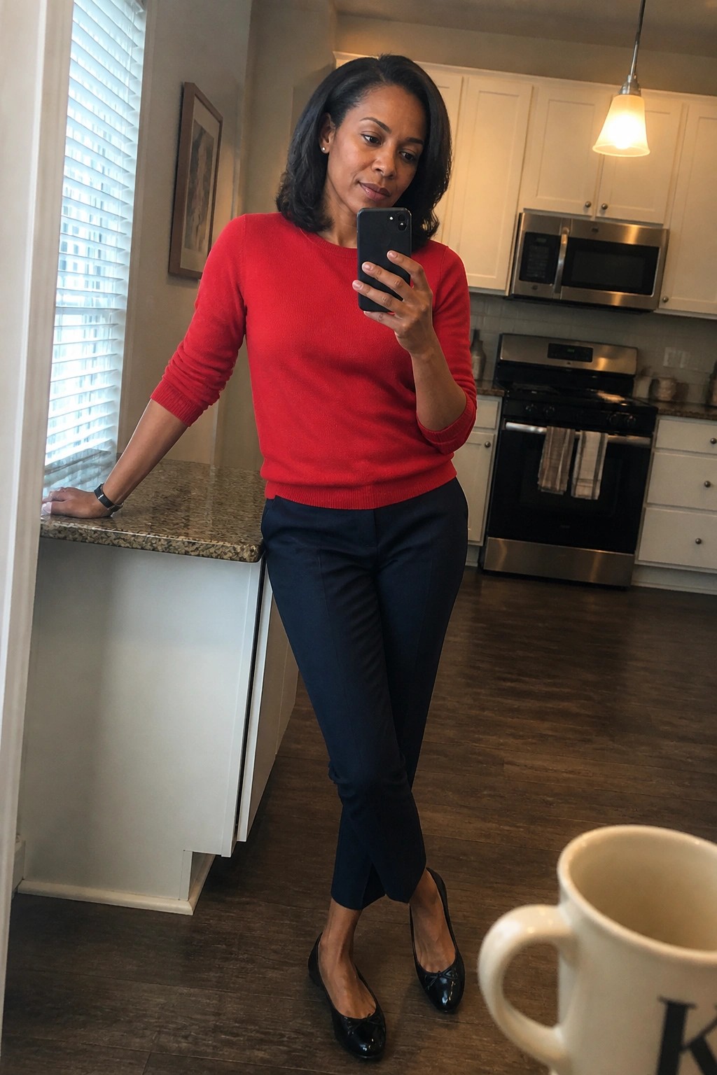 Woman in red sweater and navy pants taking mirror selfie in kitchen