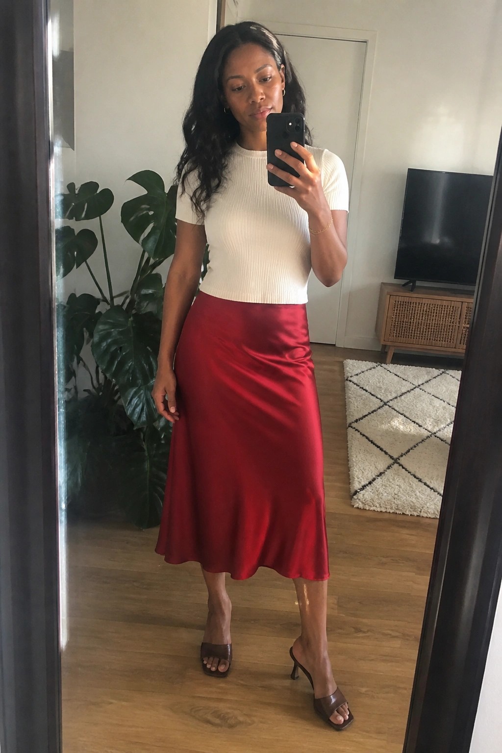 Woman in white top and red satin skirt taking mirror selfie in living room