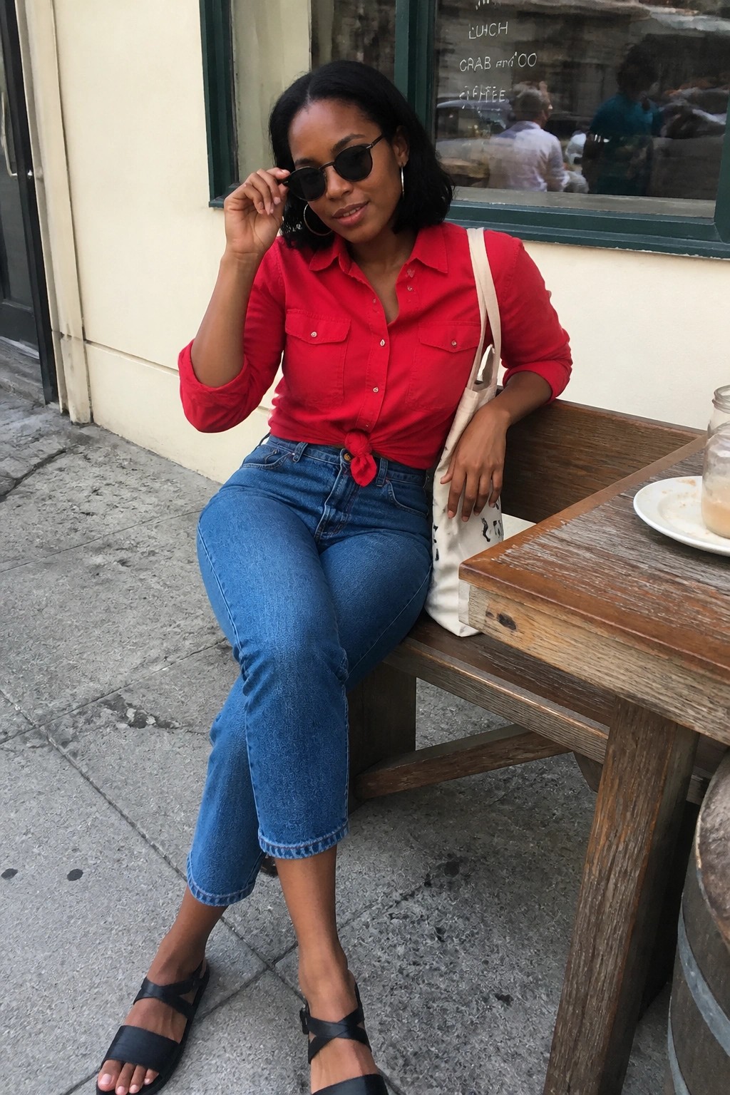 Woman in red shirt and jeans adjusts sunglasses at outdoor cafe table