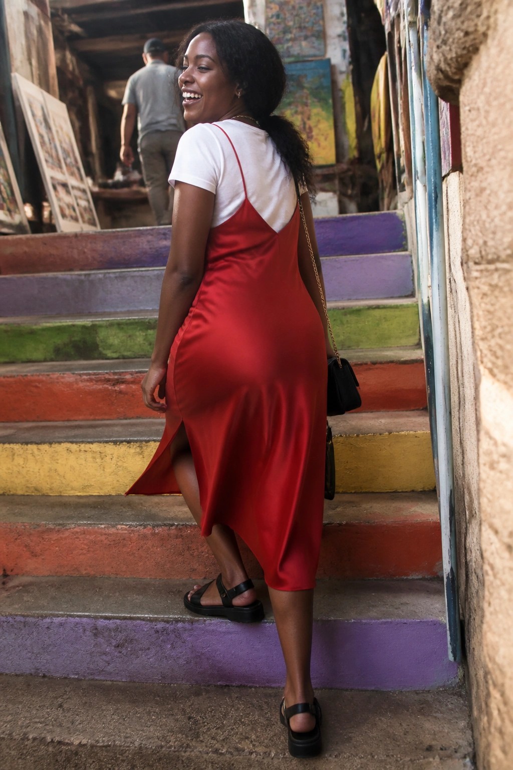 Smiling woman in red slip dress on rainbow-painted stairs with black bag