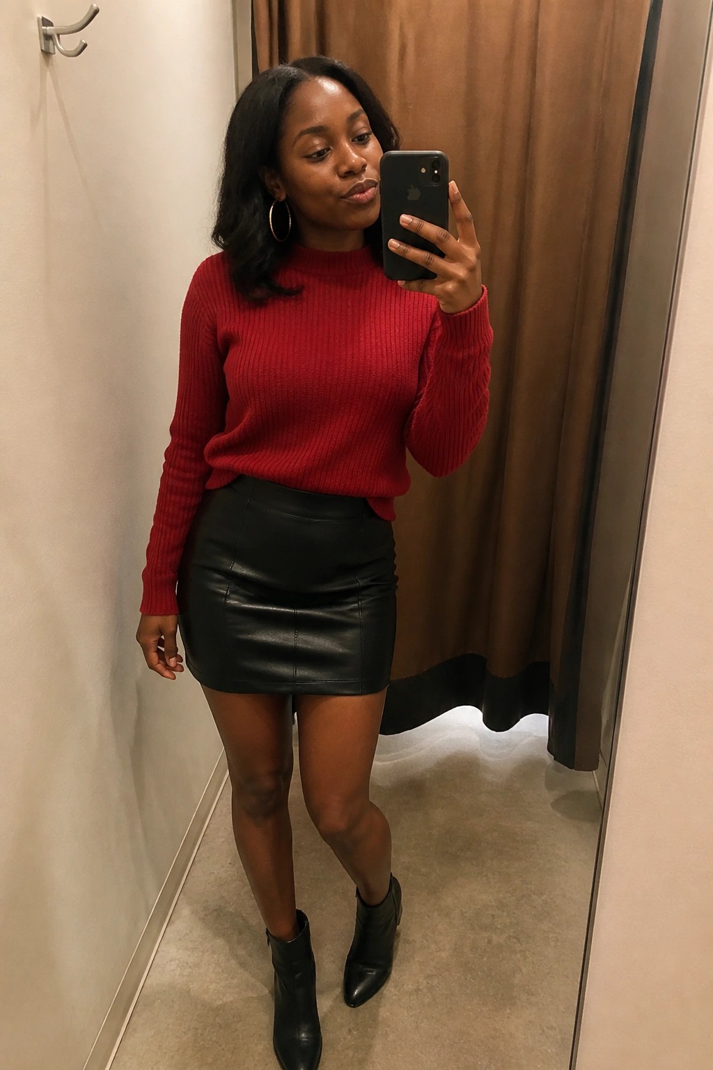 Woman in red sweater and black leather skirt taking mirror selfie in fitting room