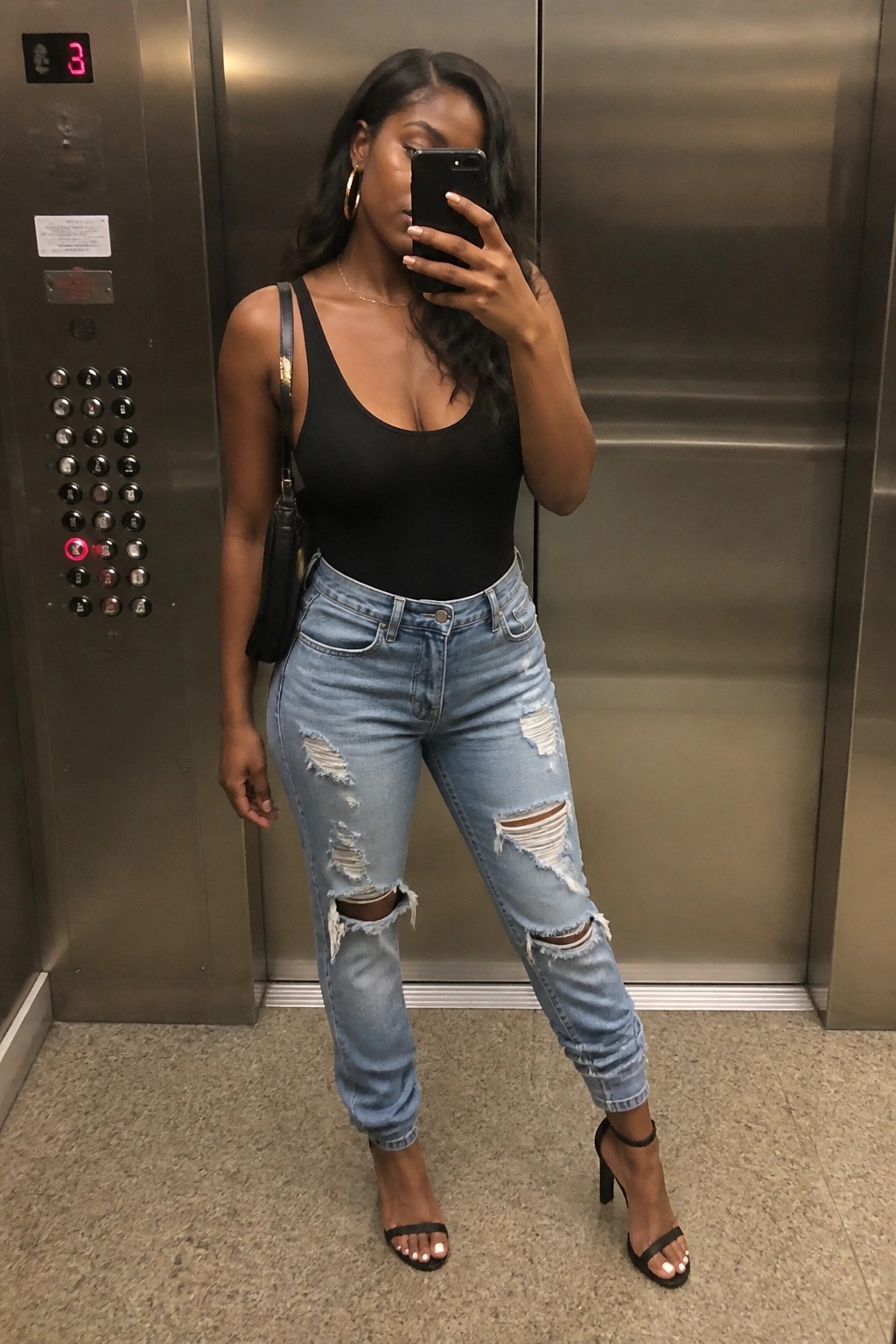 Woman in black bodysuit and ripped jeans taking mirror selfie in elevator