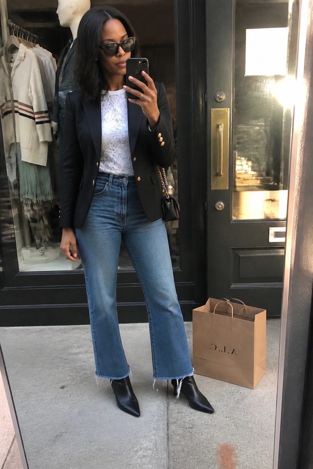 Woman in black blazer, lace top and frayed jeans taking selfie by store.