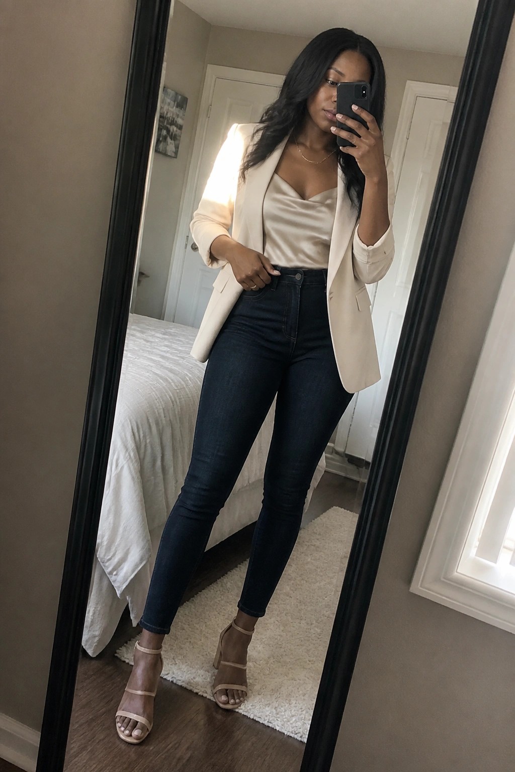 Woman in beige blazer and dark jeans taking mirror selfie in bedroom.