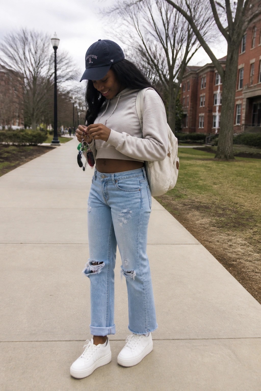 Woman wearing a cropped beige hoodie, light blue ripped straight leg jeans, white sneakers, and a navy baseball cap while holding keys on a paved walkway.